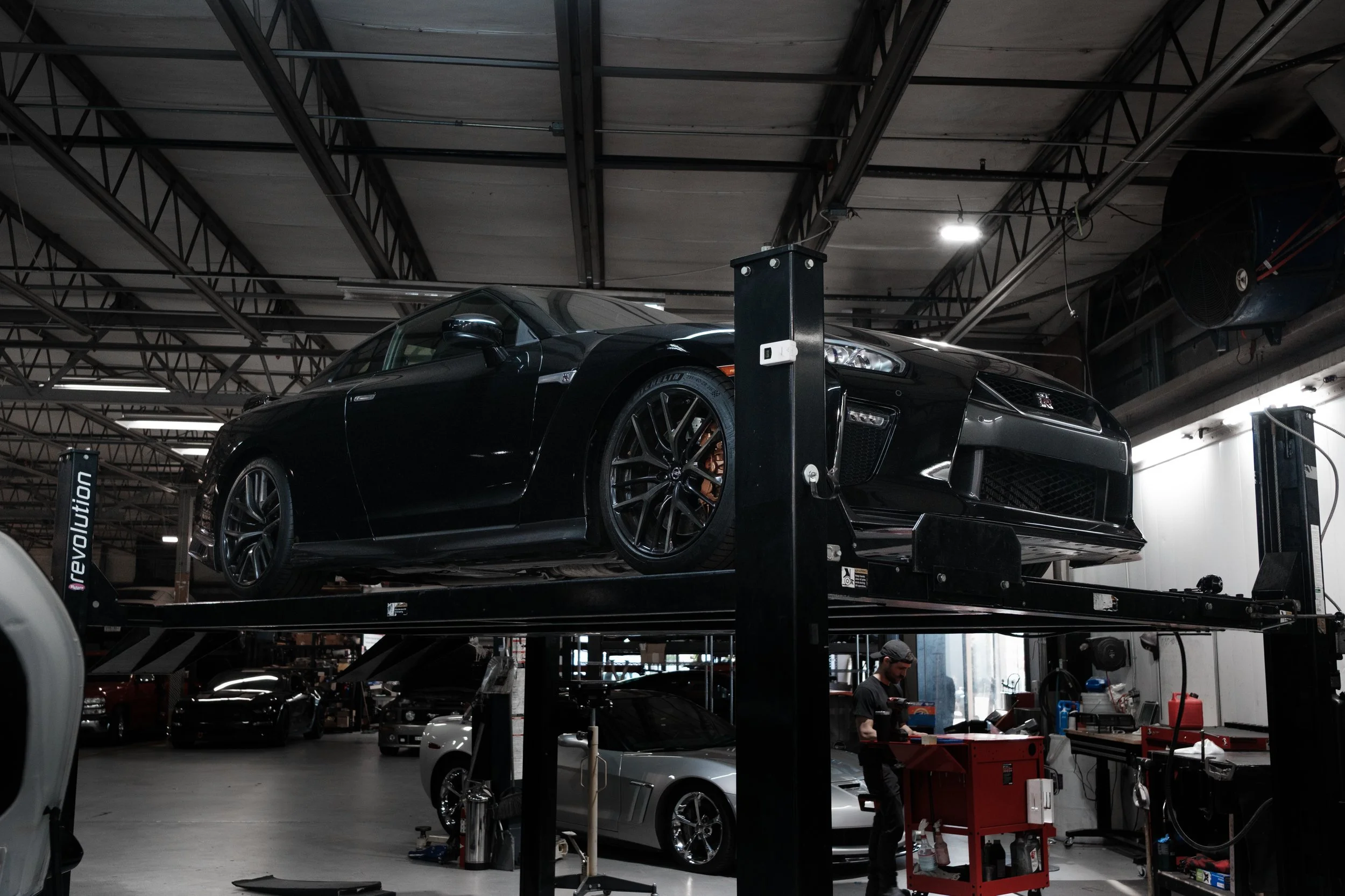 Black sports car on a lift in an auto repair shop with a mechanic working nearby.