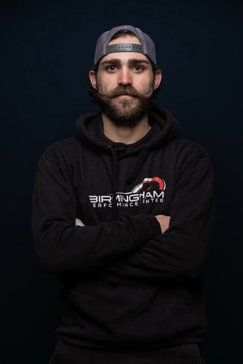 Portrait of a man with a beard and mustache wearing a backward gray baseball cap, black hoodie with logo, against a dark background.