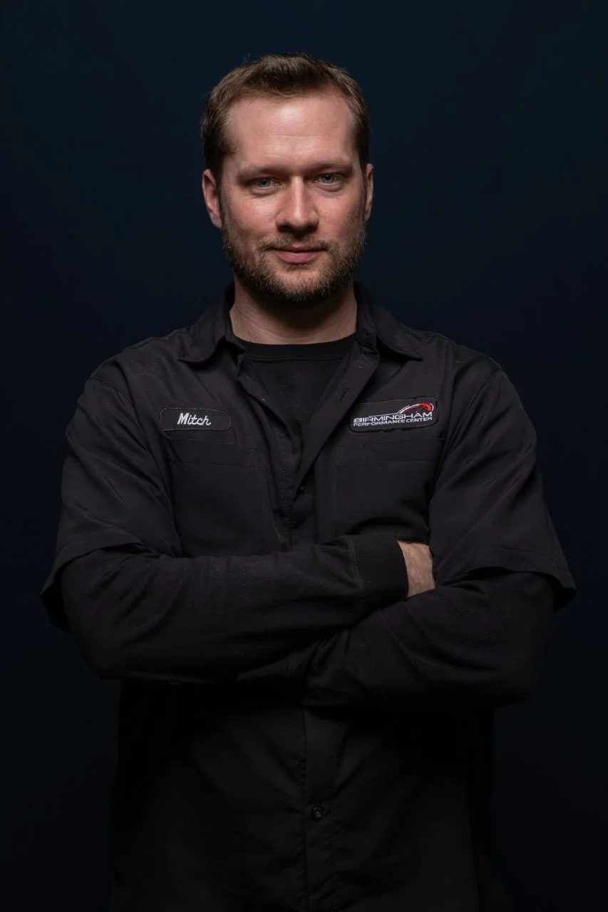 Portrait of a man with short light brown hair and a beard, wearing a black uniform with patches that say "Mitch" and "Birmingham Performance Center," standing with arms crossed against a dark background.