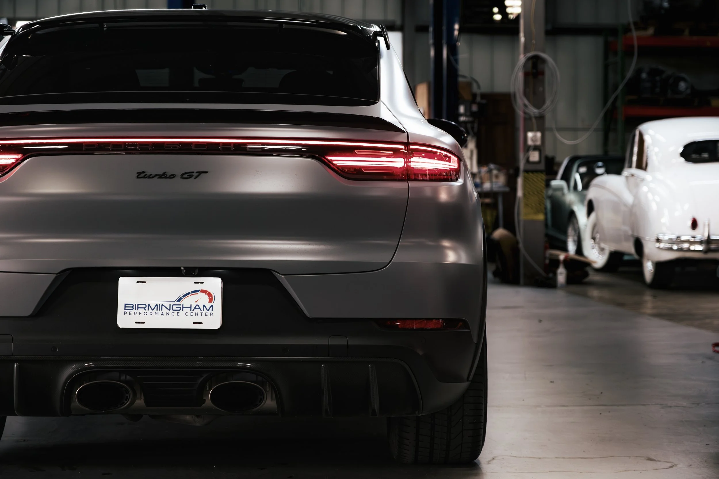 Rear view of a silver Porsche Turbo GT with a Birmingham Performance Center license plate inside a garage with other cars in the background.