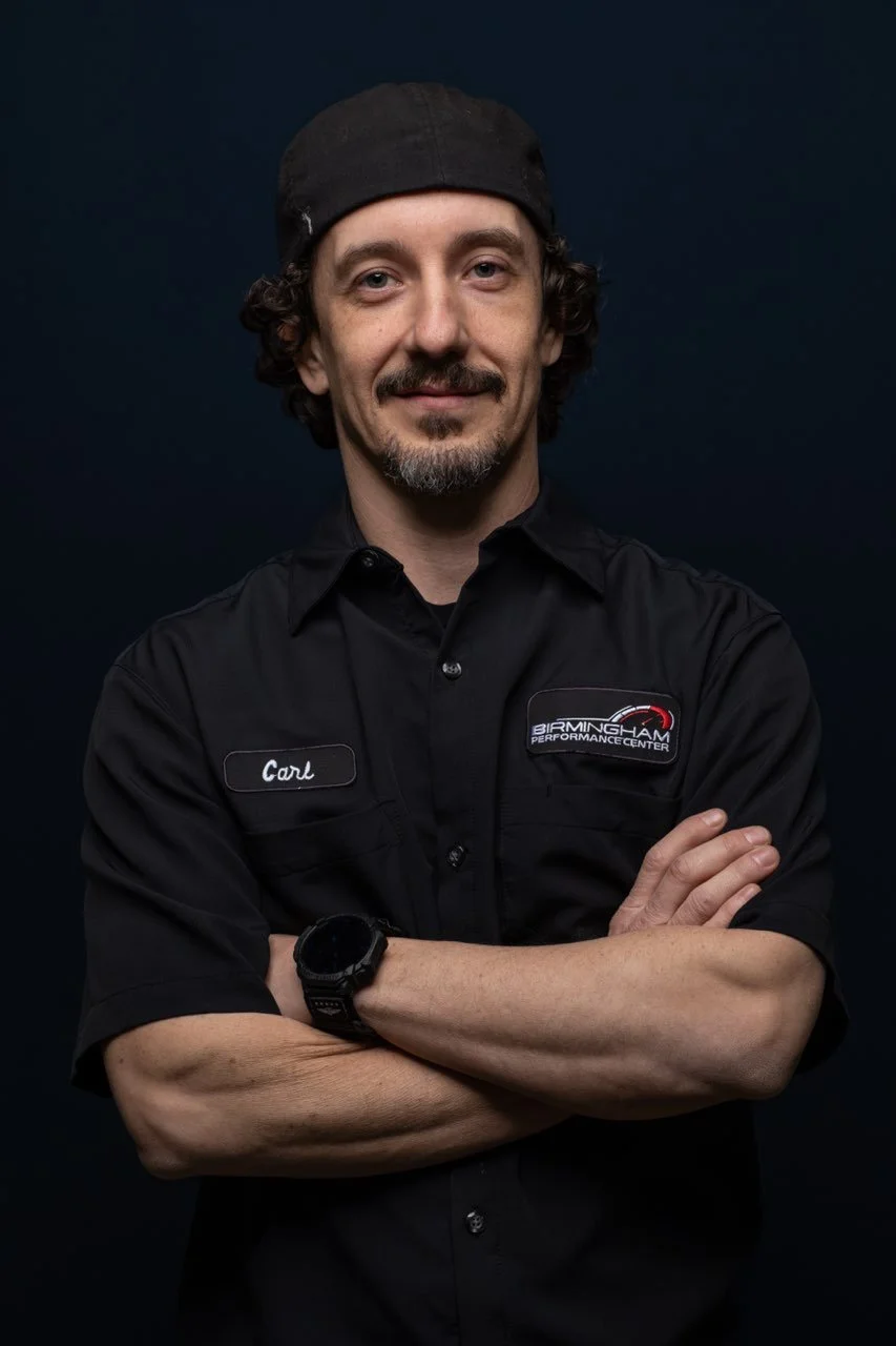 A man with curly hair, beard, and mustache wearing a black cap and black shirt with two patches, one reads 'Carl' and the other says 'Birmingham Performance Center,' posing with his arms crossed against a dark background.