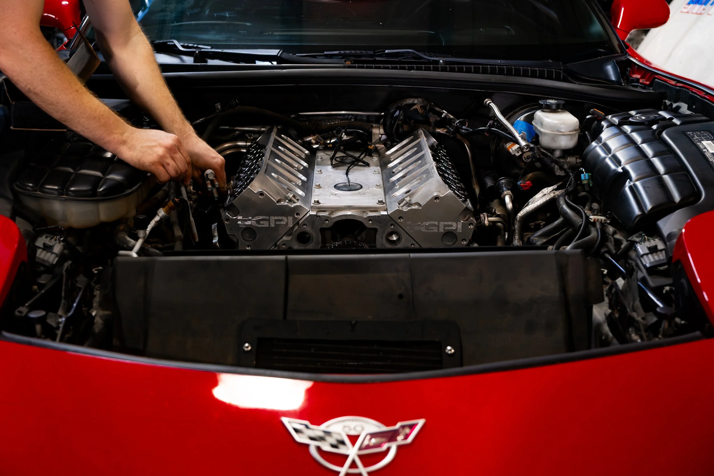 A person working on the engine of a red Chevrolet Corvette, with the hood open revealing the car's engine and various mechanical parts.