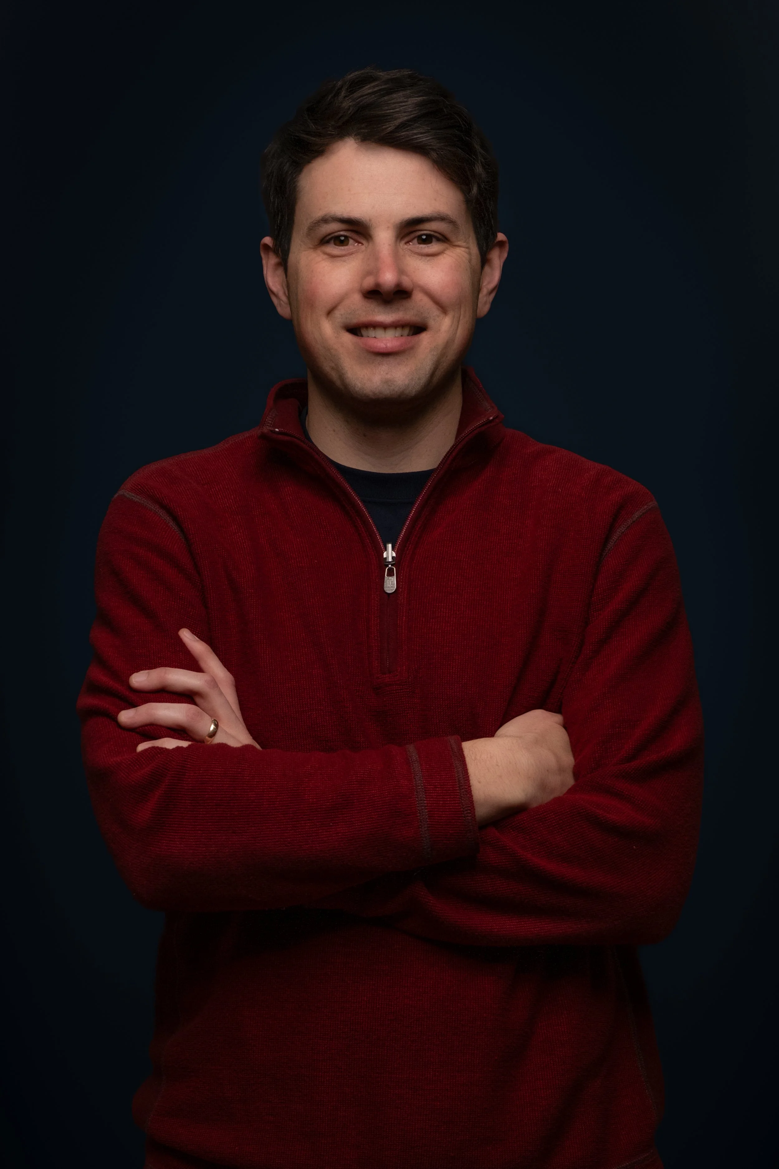 Portrait of a young man with crossed arms, smiling, wearing a red zip-up sweater against a dark background.