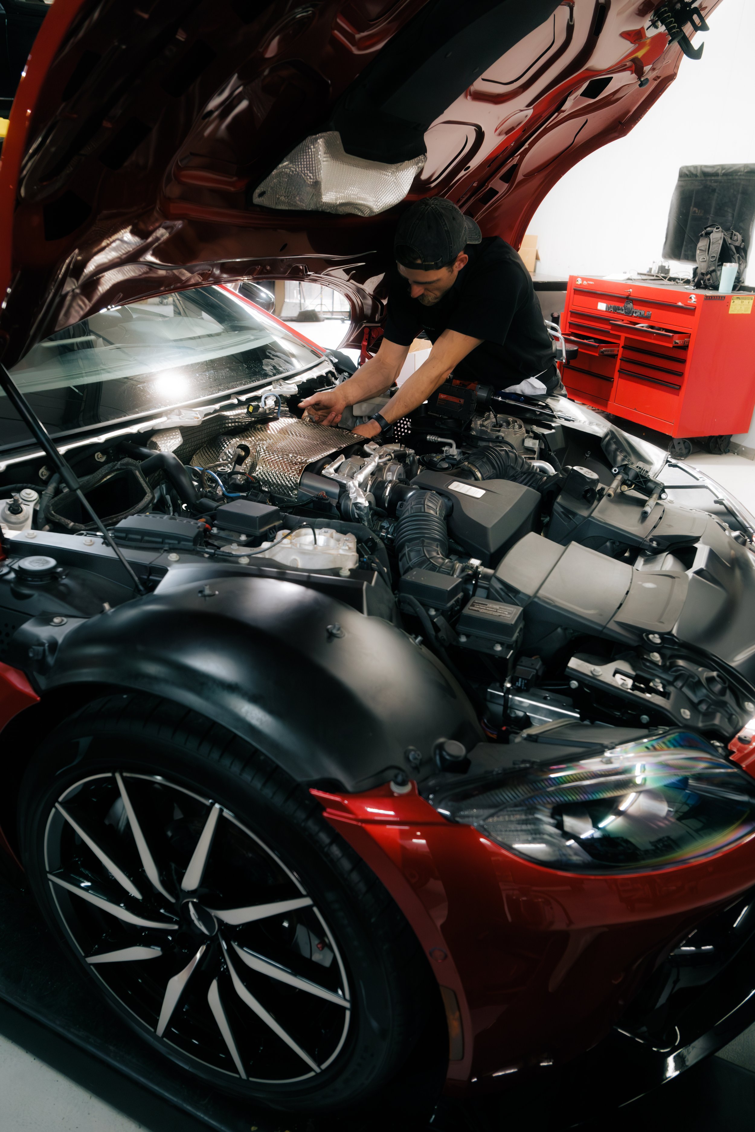 A man working on the engine of a red sports car in a garage. The car's hood is open, revealing the engine bay. The man is wearing a black shirt and a black cap, focused on maintenance or repairs. There is a red tool chest in the background.