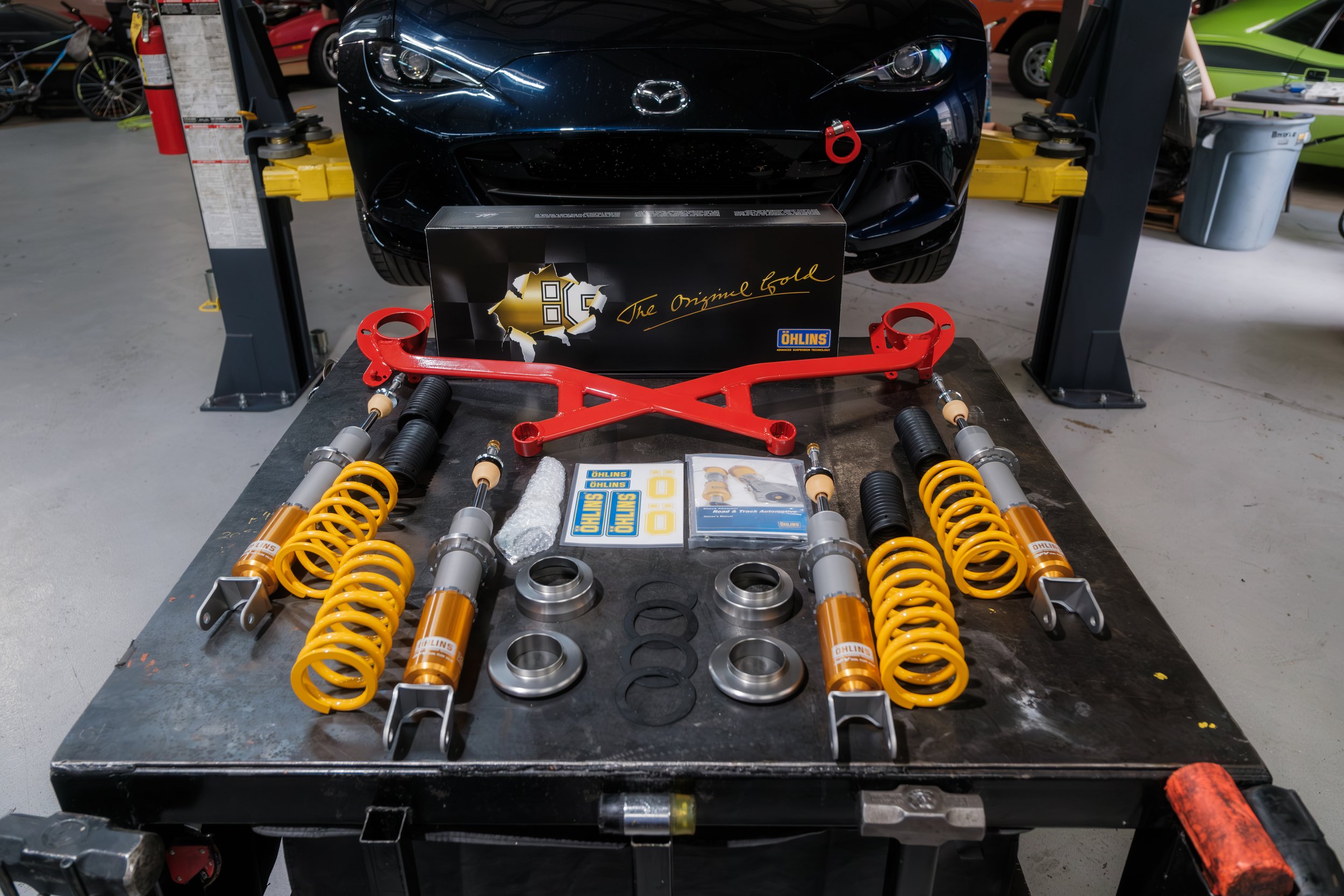 Disassembled car suspension parts, including yellow coil springs, shock absorber components, and mounting brackets, on a workbench in a garage. A black Mazda sports car is raised on a lift in the background.
