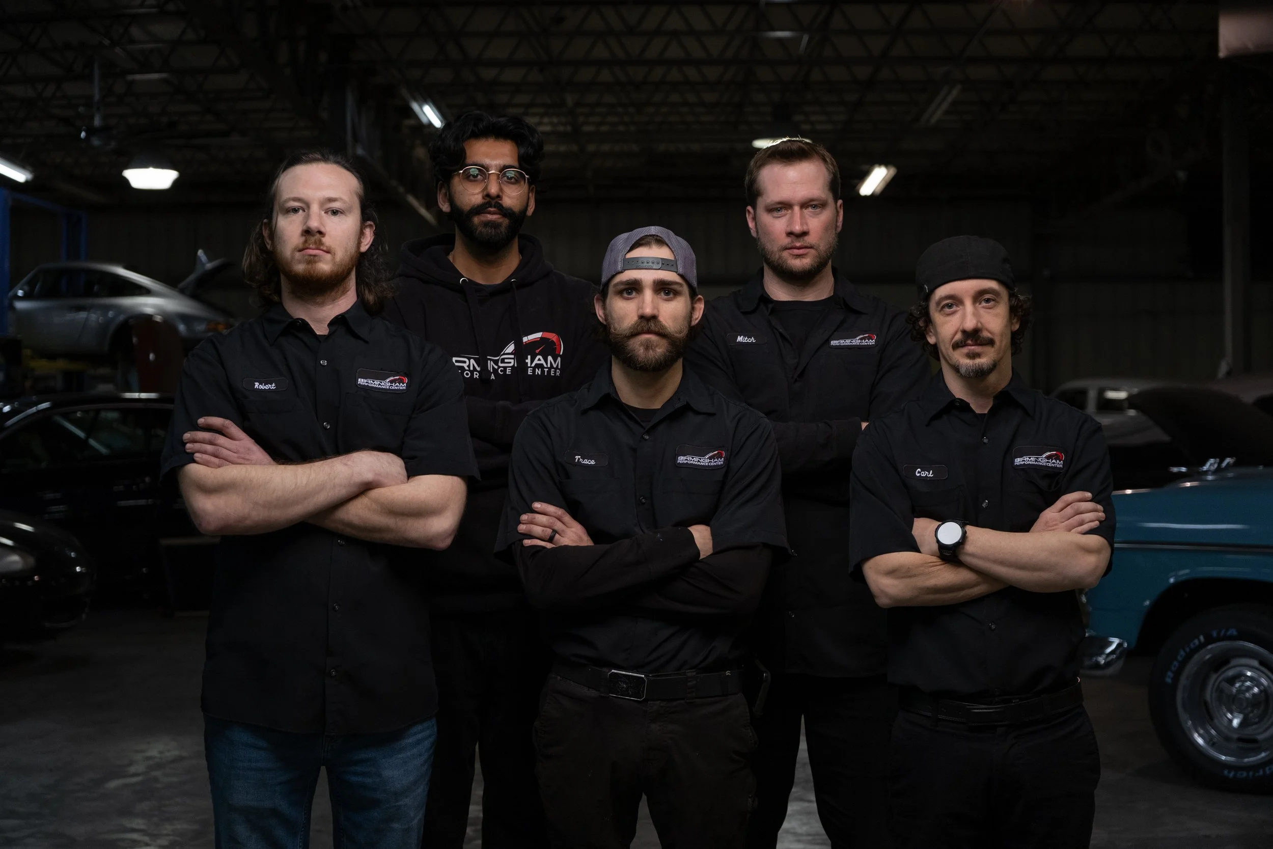 Mechanics and business owner in black shirts with embroidered Birmingham Performance logos, standing with arms crossed inside a garage with cars, some elevated on lifts, in the background.