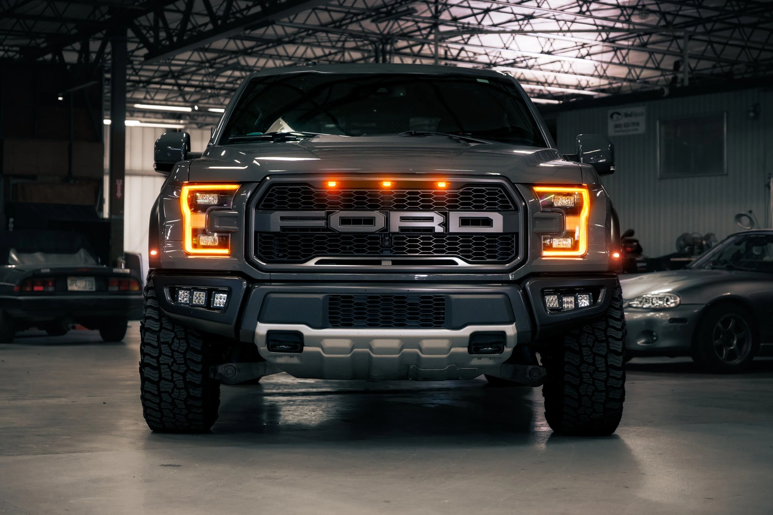 Front view of a black Ford pickup truck with illuminated headlights and grille, parked indoors with other vehicles in the background.