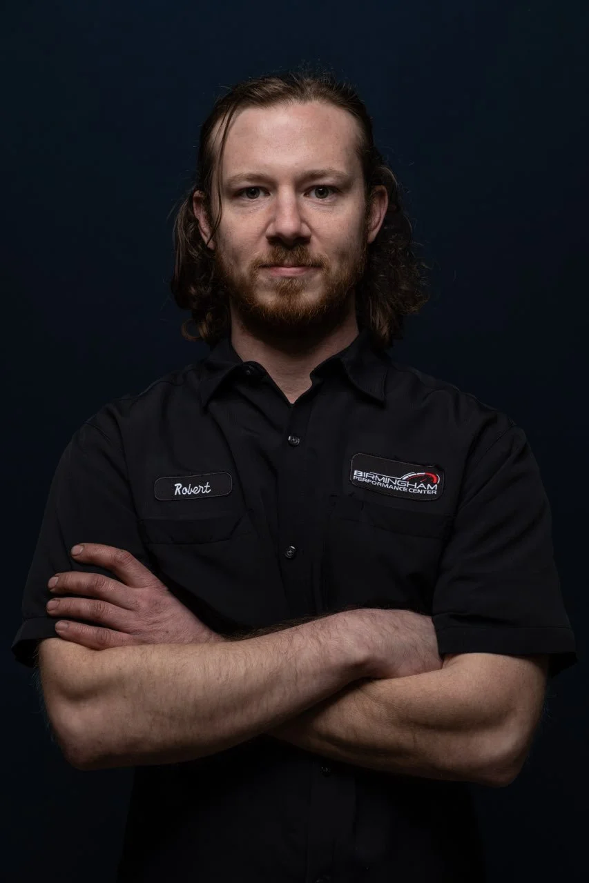 A man with long hair and a beard wearing a black shirt with a name tag 'Robert' and a Birmingham Performance Center logo, standing with crossed arms against a dark background.