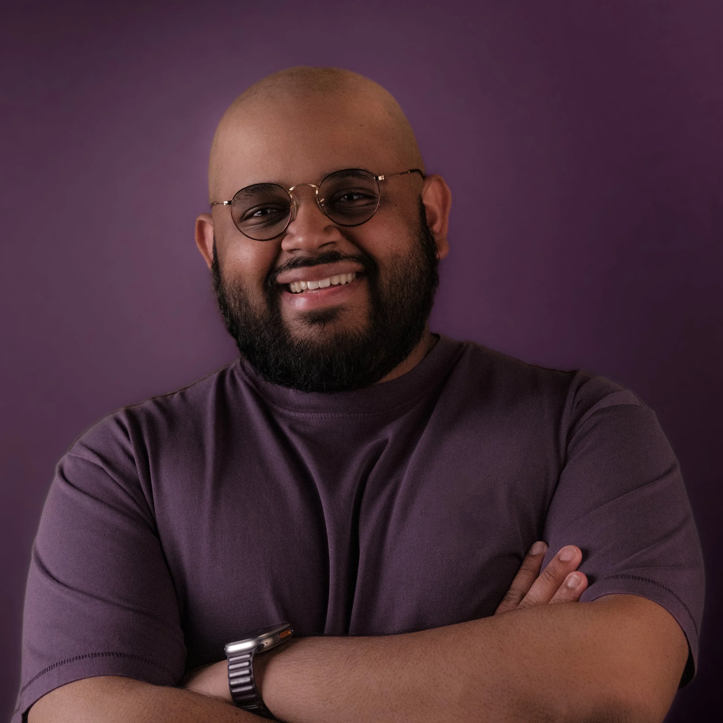 A smiling man with glasses, bald head, and beard crossing his arms against a purple background.