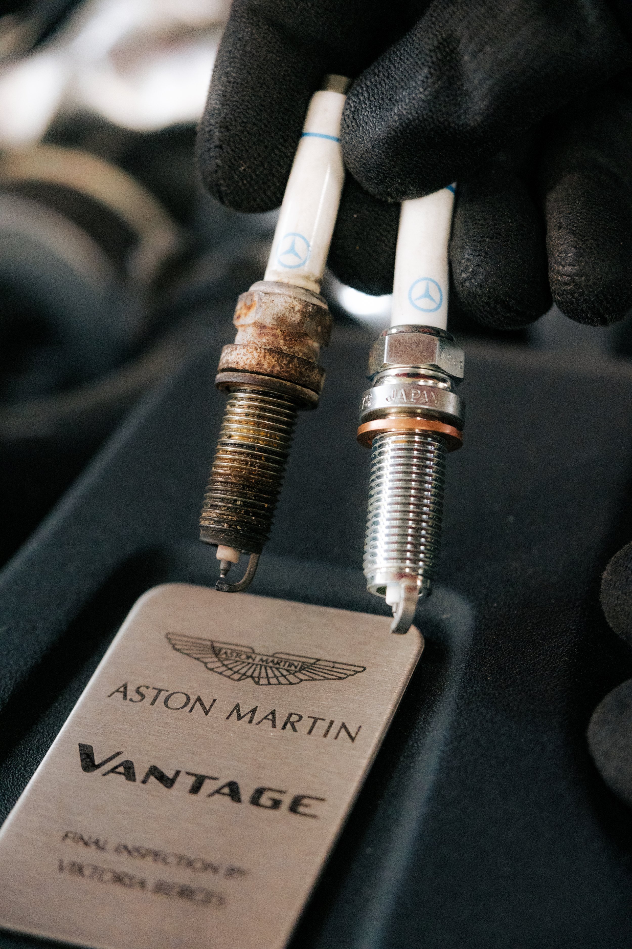 Comparison of a rusty and a new Mercedes-Benz spark plug with an Aston Martin Vantage age card.