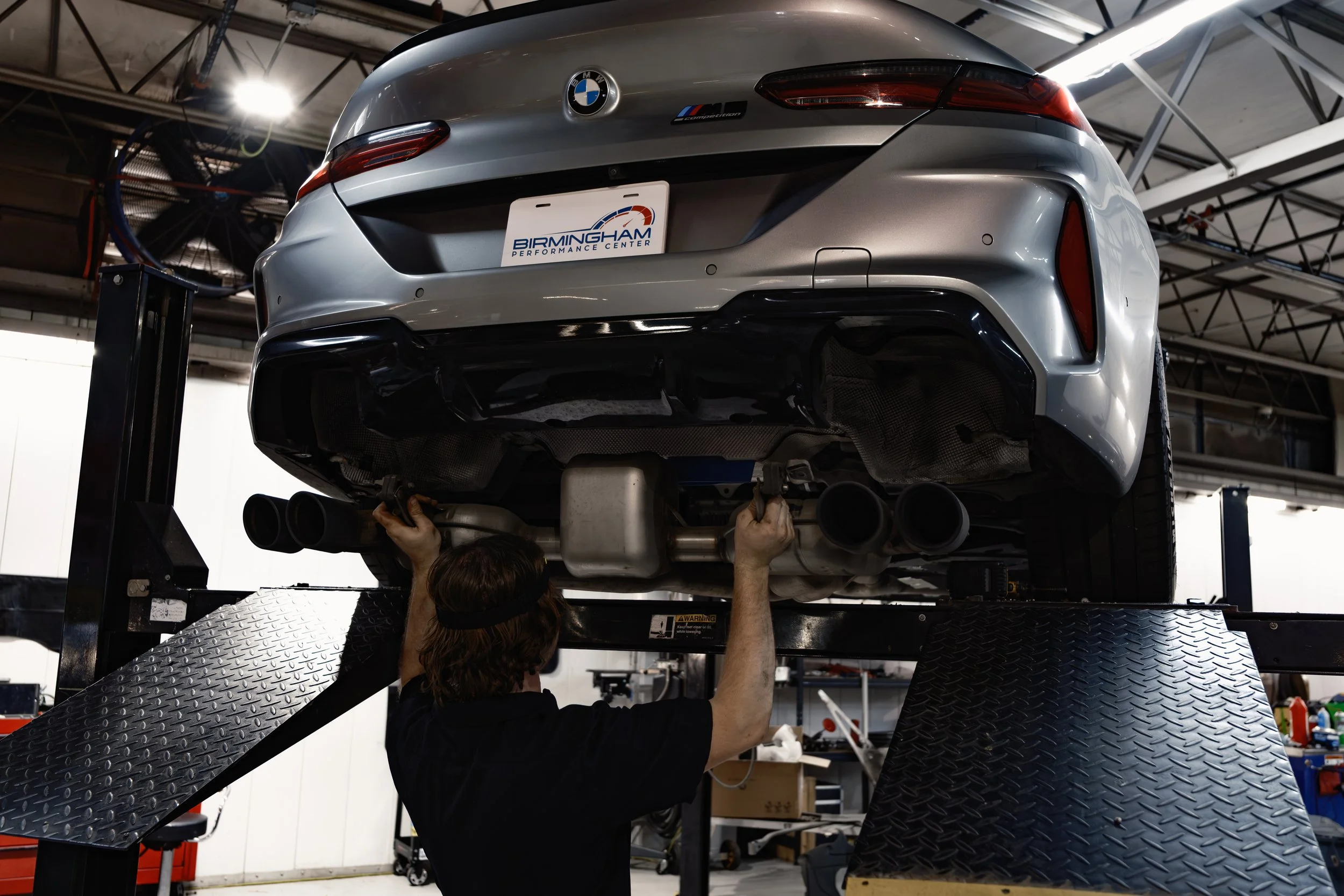 A mechanic working under a raised BMW sports car in an auto repair shop, inspecting the exhaust system.