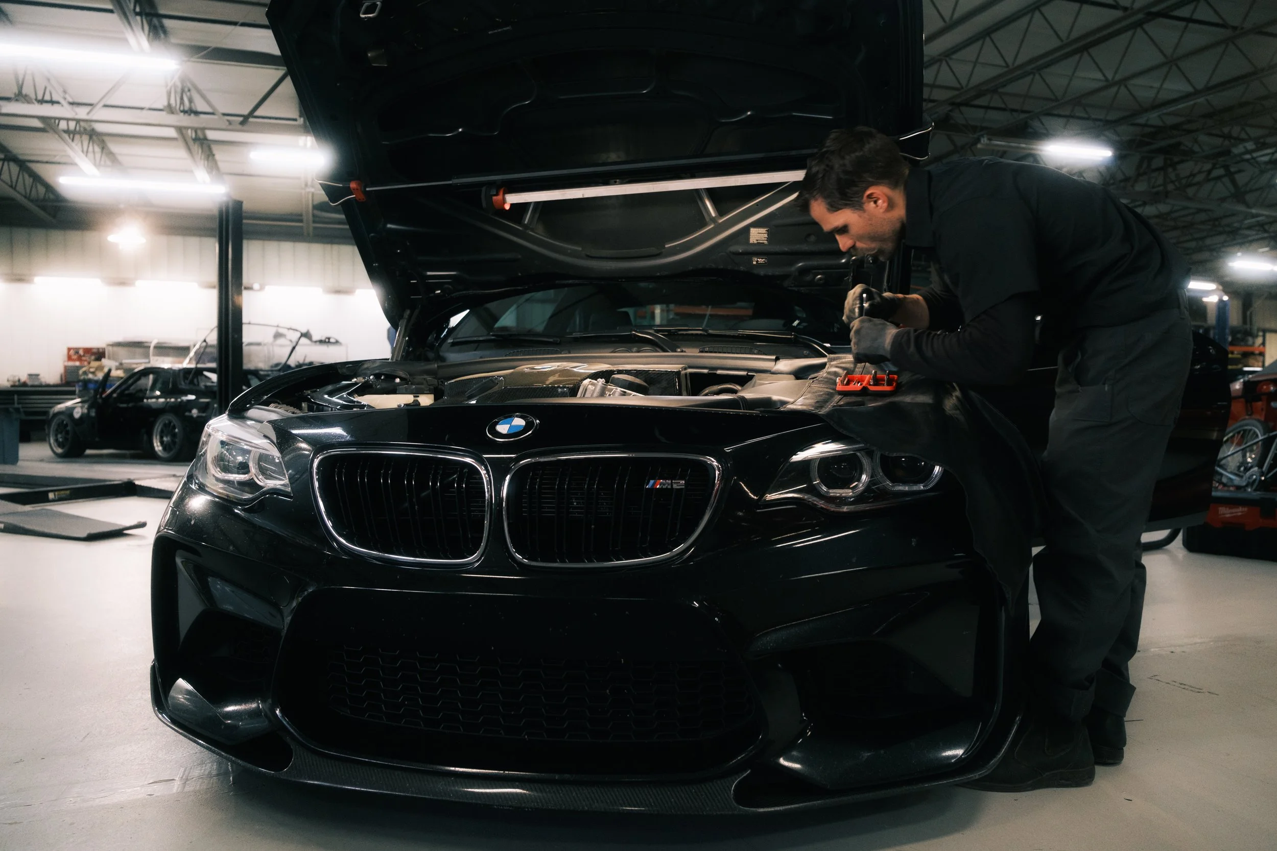 Mechanic working on the engine of a black BMW M series car inside a workshop.