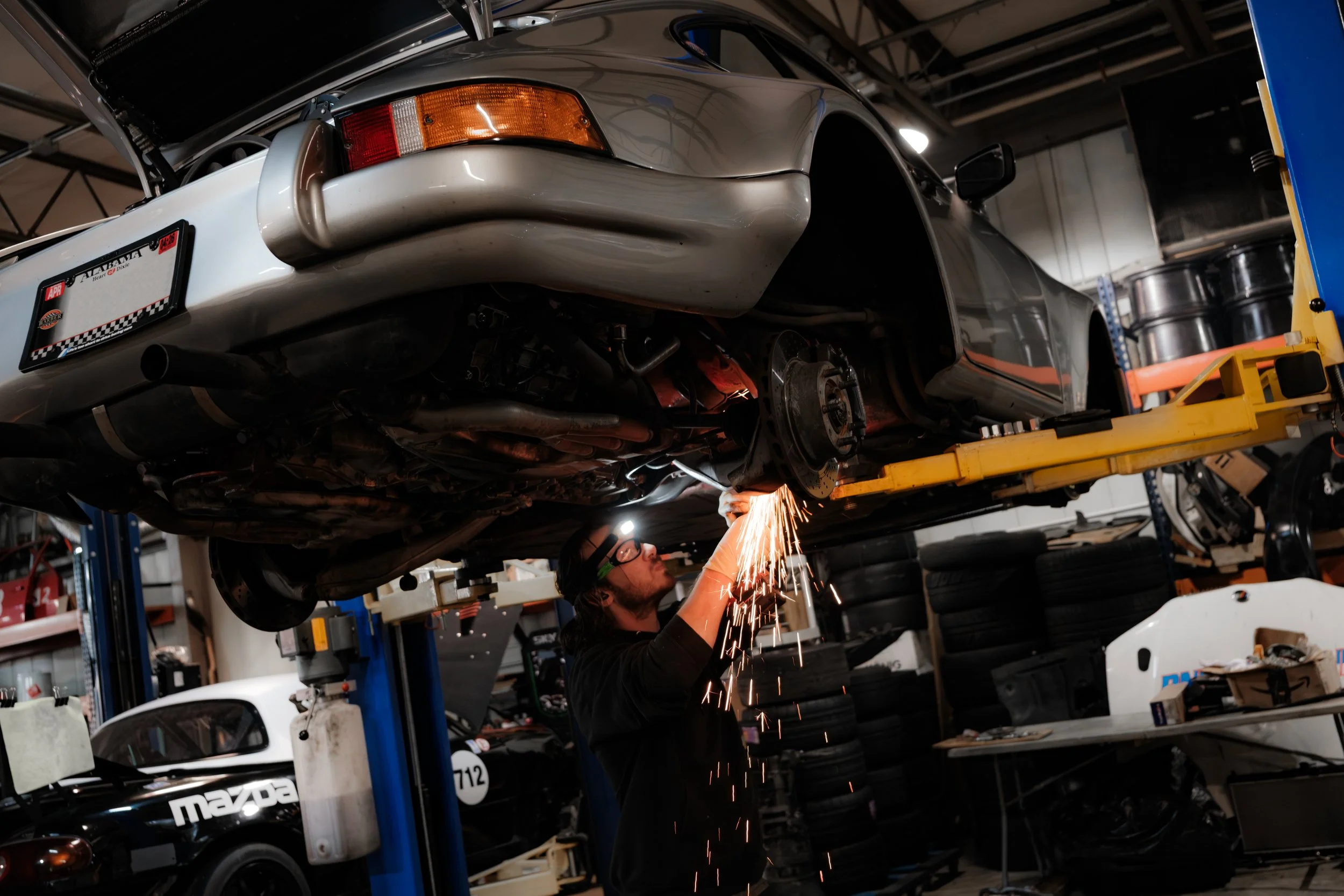 A mechanic working under a car elevated on a lift, welding or grinding near the rear wheel area in an auto repair shop.