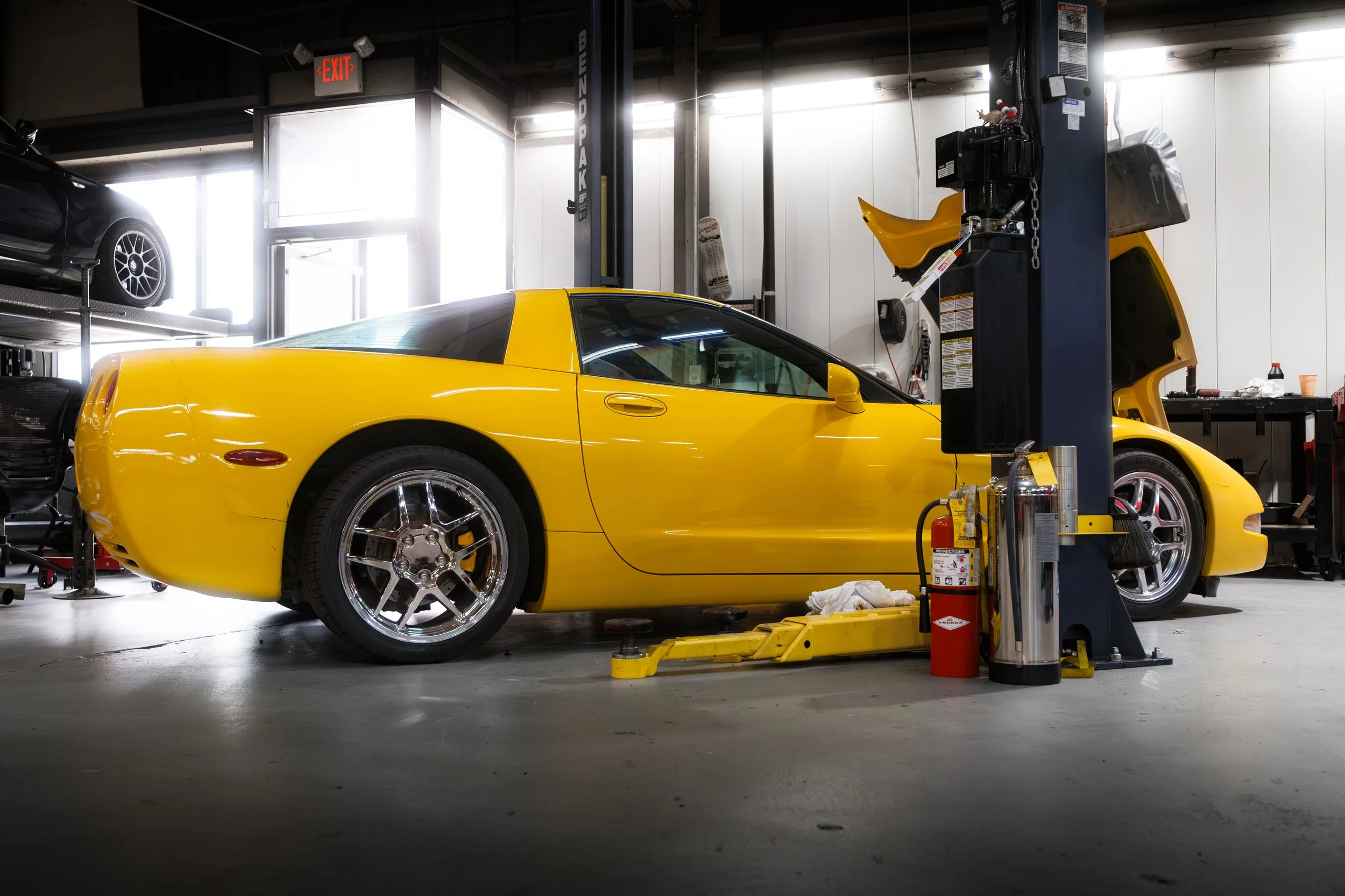 A yellow sports car is inside an auto repair shop, raised on a hydraulic lift with the front hood open, surrounded by tools and equipment.