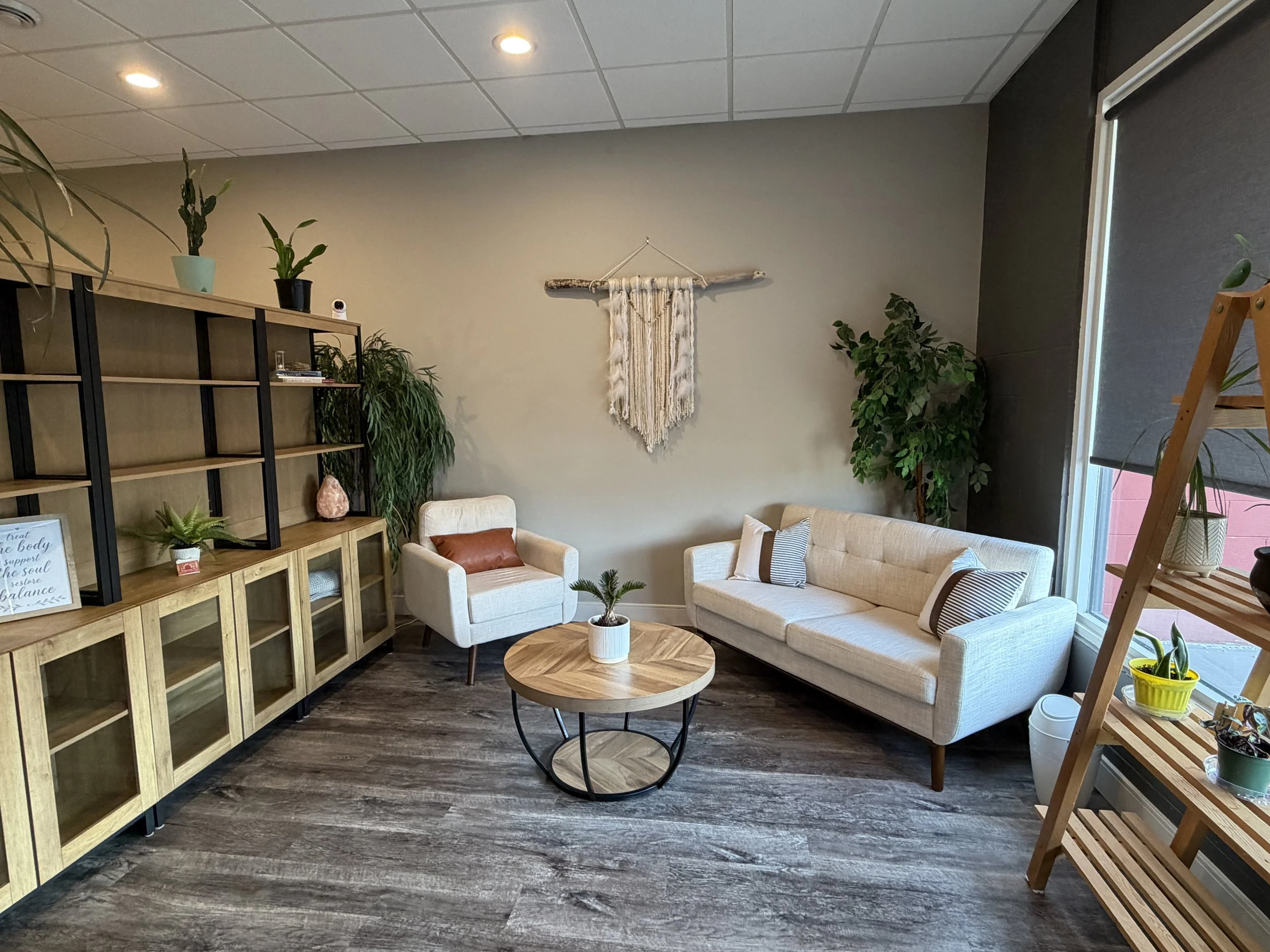 A cozy living room with a beige sofa and matching armchair, a round wooden coffee table with a white potted plant, assorted potted plants on a wooden shelf, wall decor including a woven hanging, and large windows with shades, accented by plants and wooden furniture.