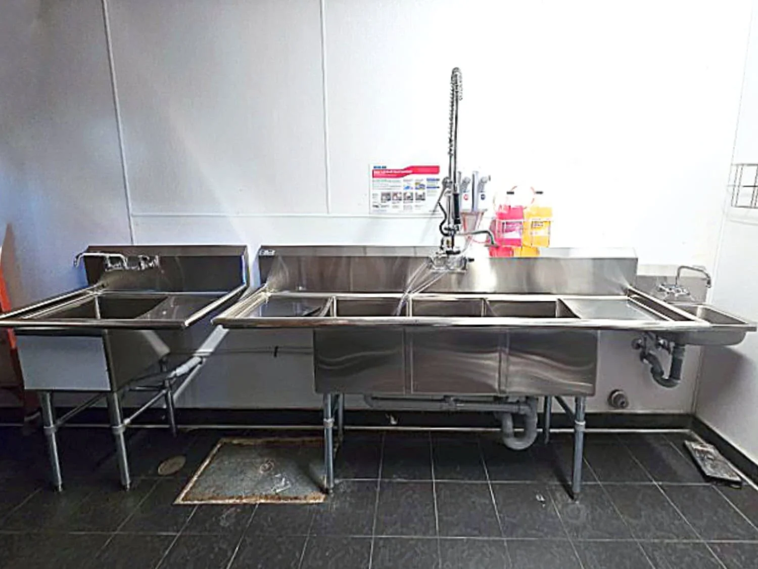 Stainless steel industrial sink with multiple basins, a high faucet, and drainage pipes underneath, located in a commercial kitchen or food preparation area.
