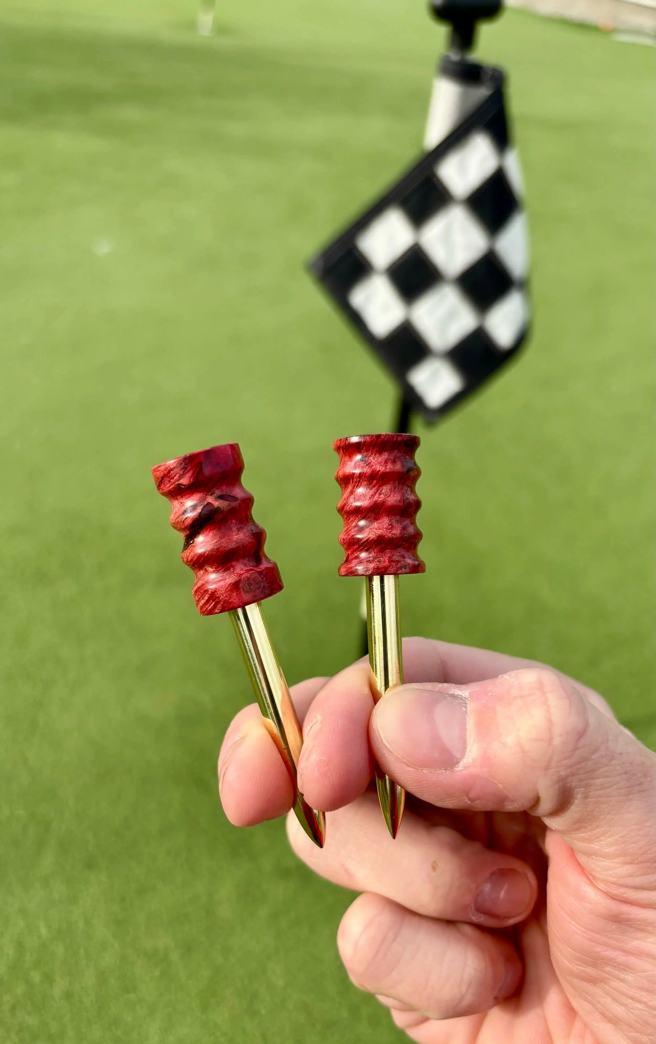 A person holding two golf tee markers with red tops, with a golf flag displaying a black and white checkered pattern in the background on a green golf course.