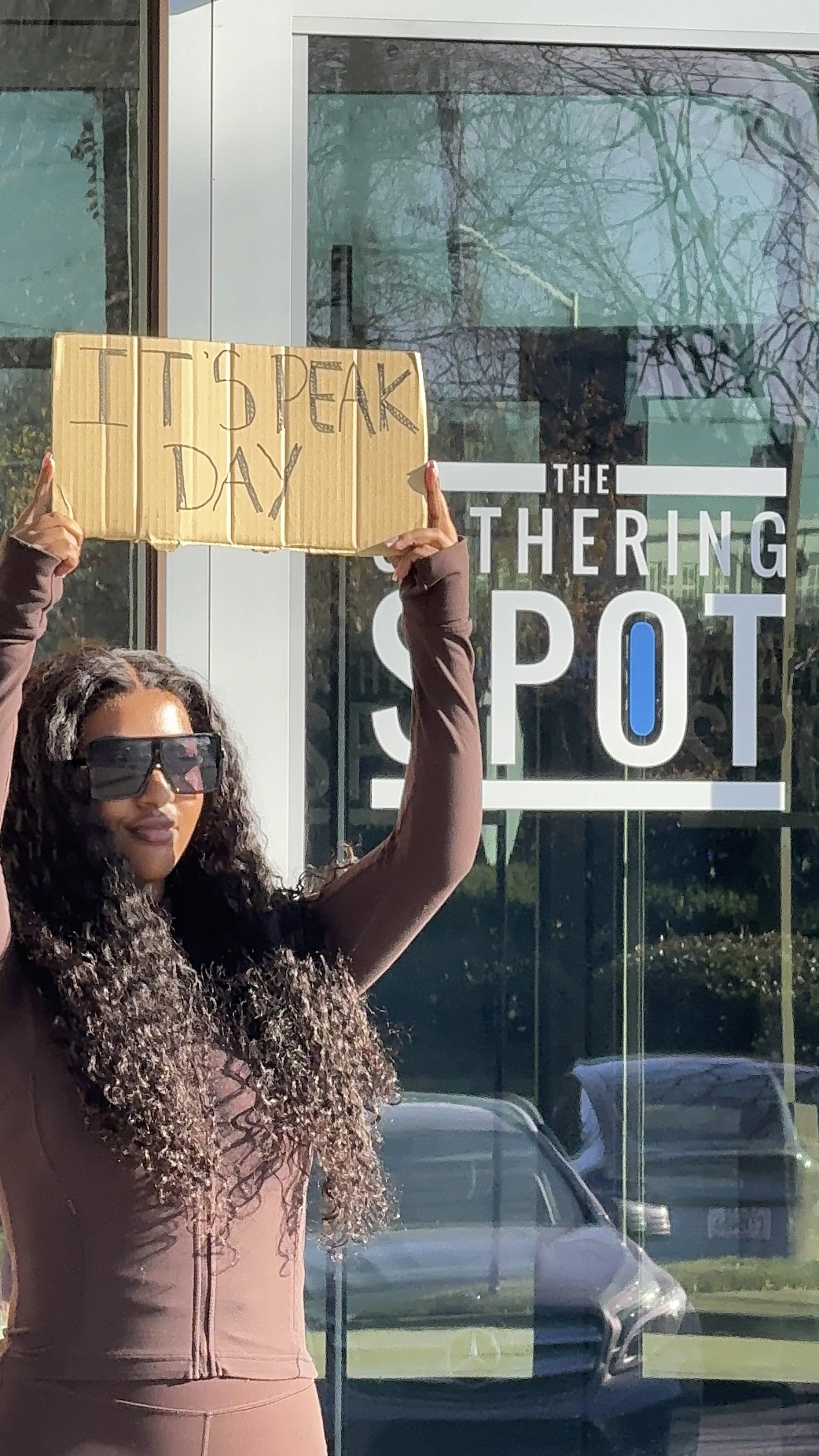 A woman with long curly hair and sunglasses holding a cardboard sign that reads 'I' and 'DAY' with her arms raised. She is standing in front of a glass window with text that reads 'THE SHERING SPOT'.
