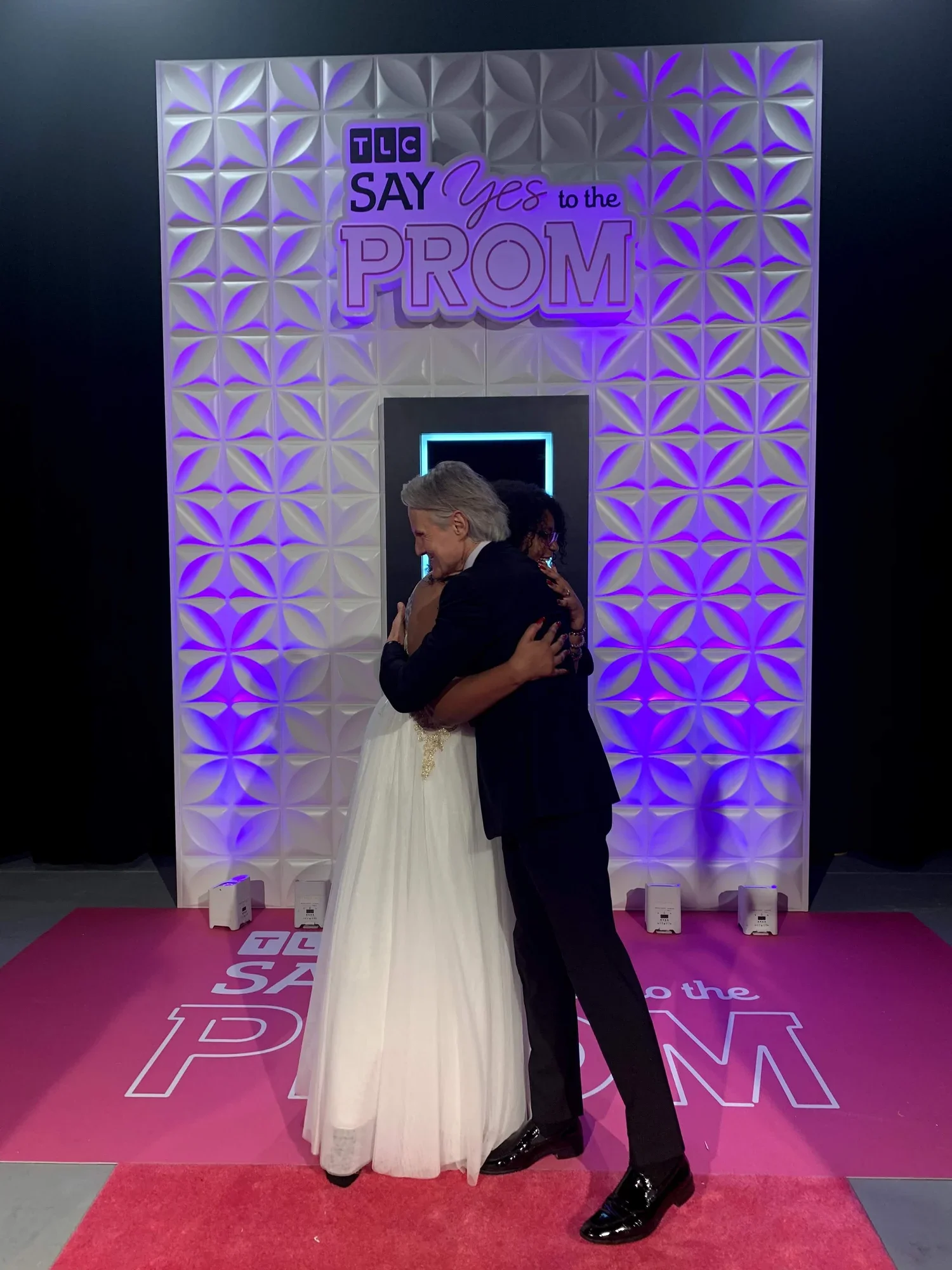 A couple hugging on a pink carpet at a prom-themed event with a sign that says "TLC SAY Yes to the PROM;" the background features a decorative wall with purple lighting.