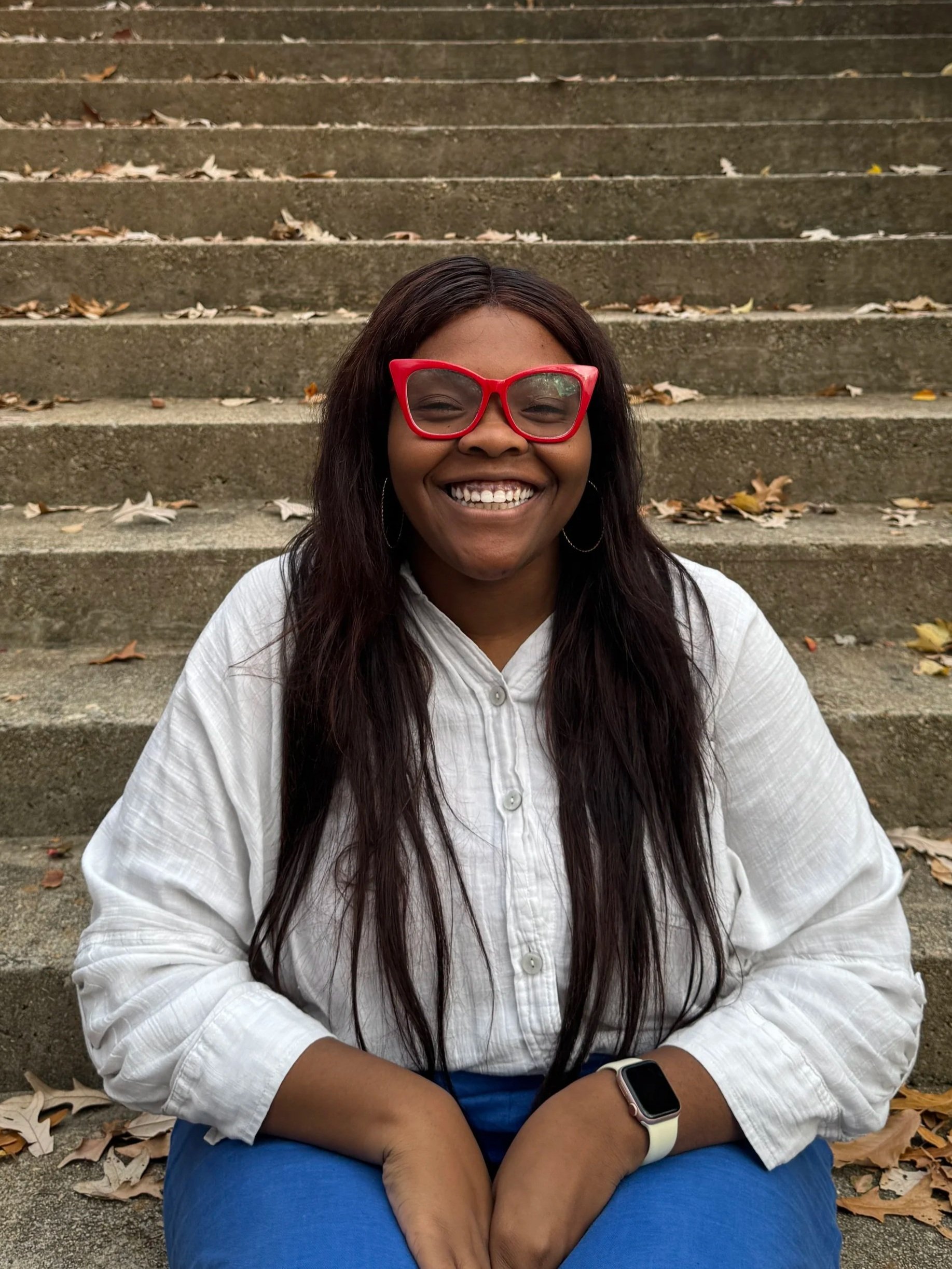 A woman smiling and sitting on outdoor stairs with fallen leaves, wearing red glasses, a white shirt, blue pants, and a smartwatch.