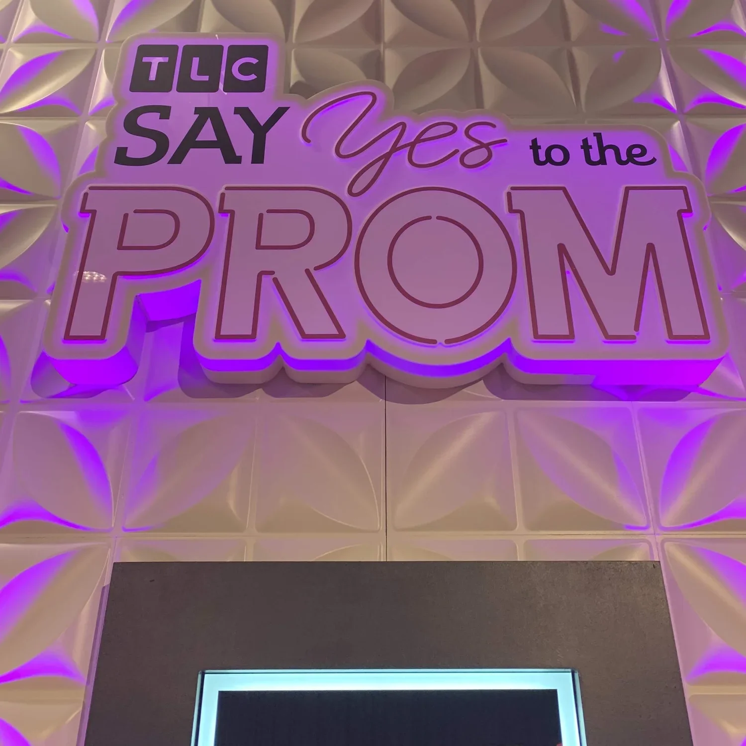 Neon sign on a wall that says, 'TLC SAY Yes to the PROM' with purple and white lighting.