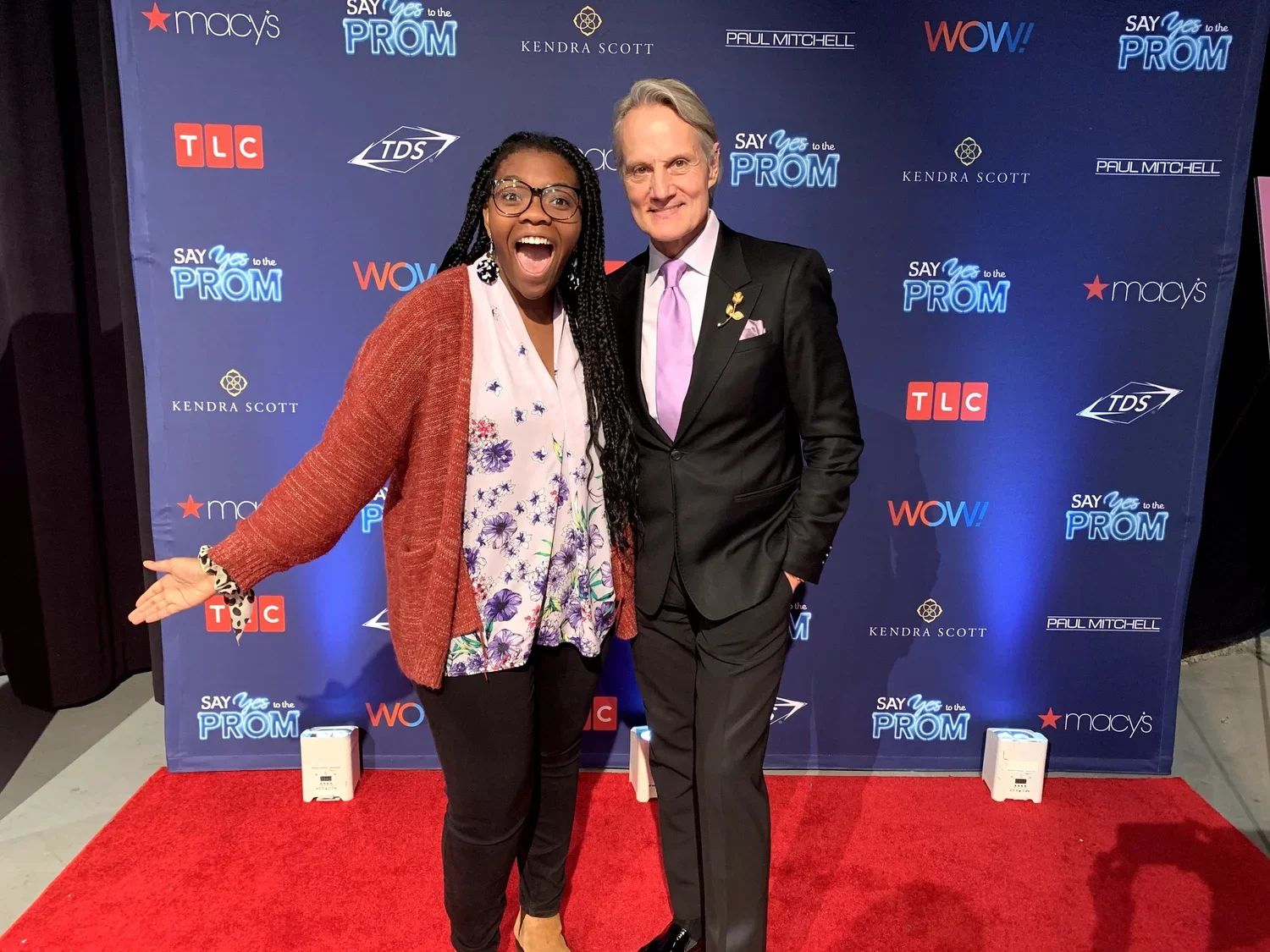 A woman and a man posing happily together on a red carpet at a promotional event, with a blue backdrop featuring logos for Macy's, TLC, TDS, WOW, and Kendra Scott. The woman is wearing glasses, a colorful floral blouse, a red cardigan, and black pants. The man is dressed in a black suit and a light pink tie.