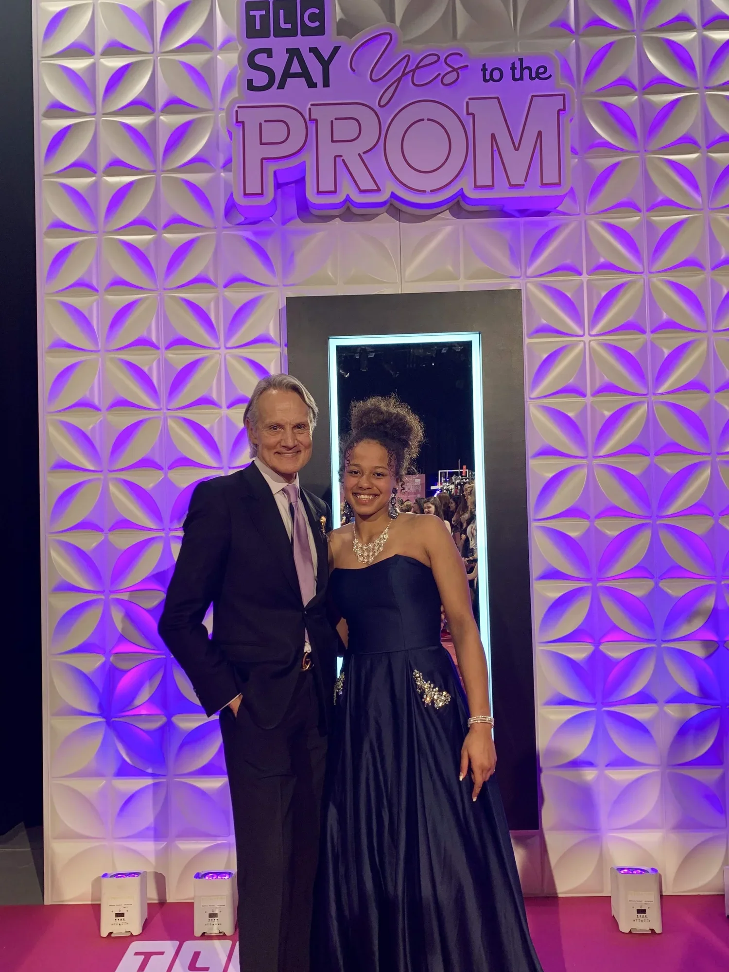 Two people, a man and a woman, dressed in formal attire, standing in front of a decorative wall with a sign that says ‘TLC SAY Yes to the PROM’ in purple and pink letters. The man is wearing a black tuxedo and purple tie, and the woman is wearing a strapless black ball gown with jewelry. They are smiling at the camera, with a mirror reflecting other people and a makeup station in the background.