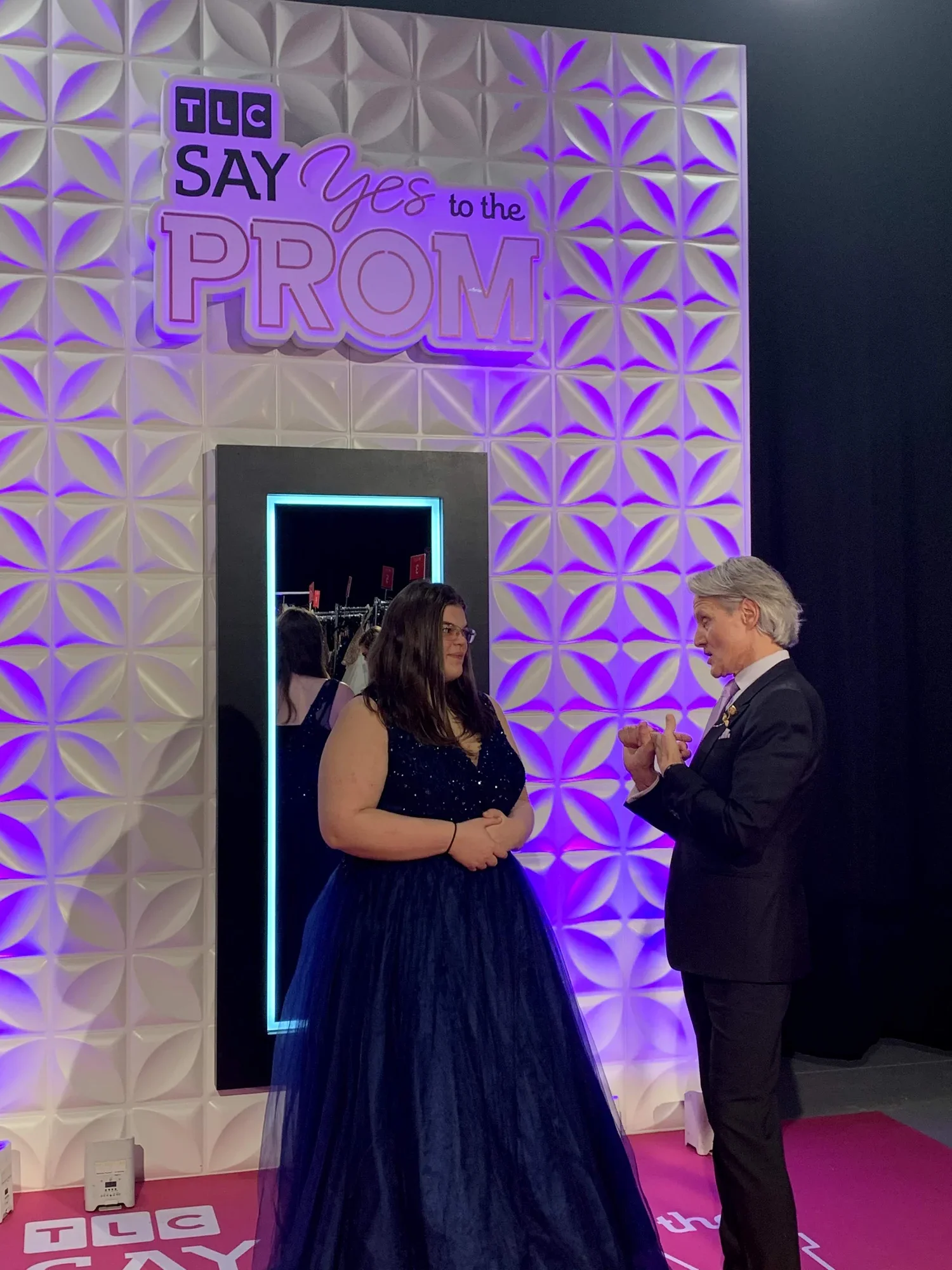 A woman in a navy blue strapless gown and a man in a dark suit are talking at an event with a decorative white wall lit with purple lights in the background. The wall has a sign that reads "TLC Say Yes to the PROM" and a bright rectangular mirror with a neon light border.