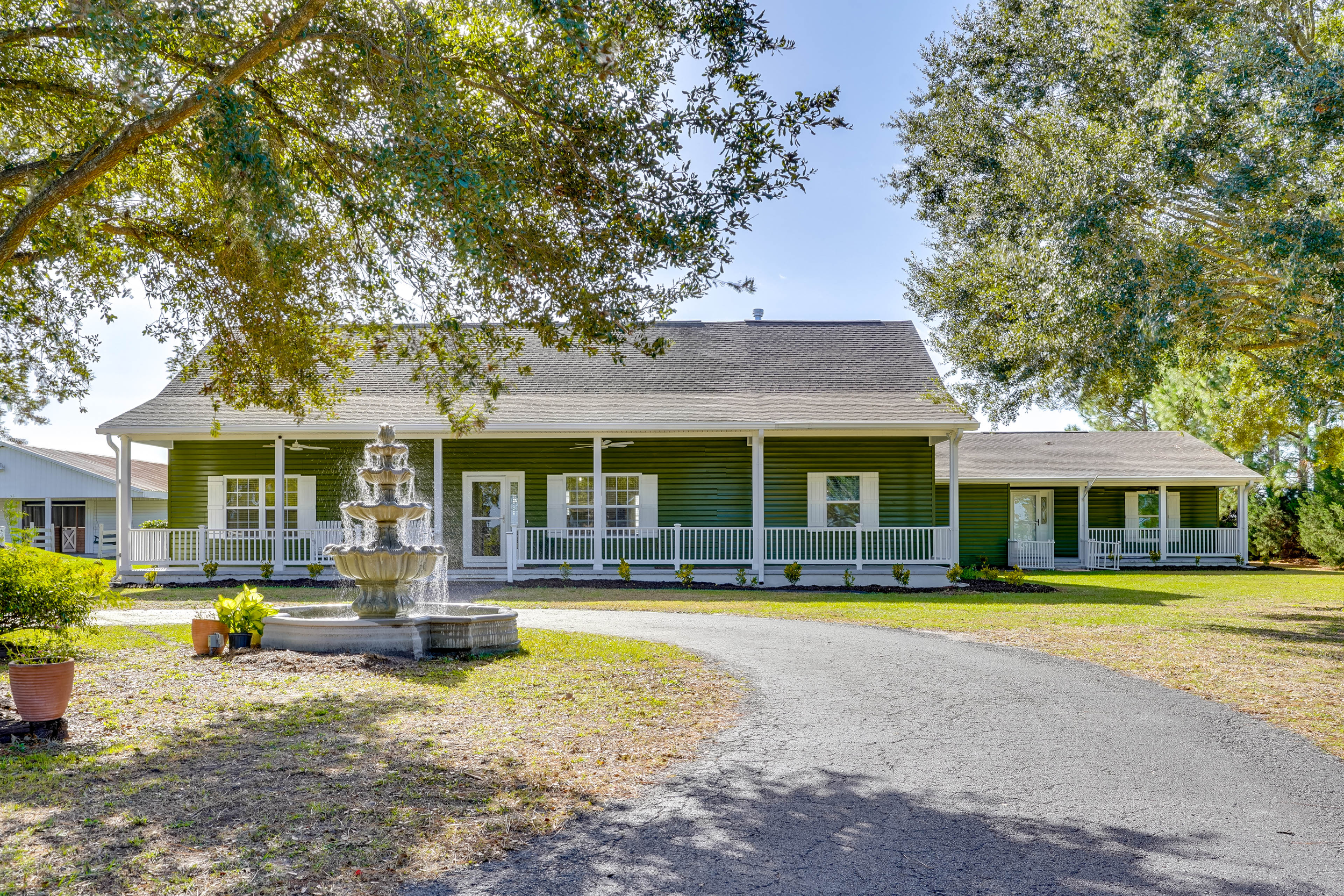 A green house with a covered porch, surrounded by trees, with a fountain in the front yard and a curved driveway.