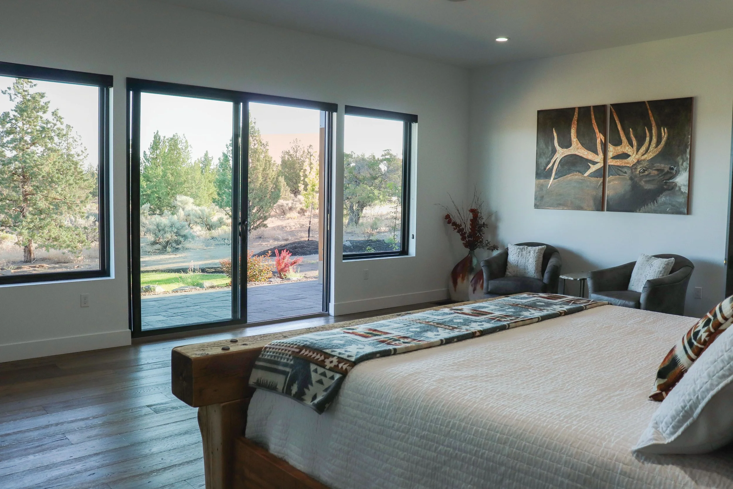 A bedroom with a large wooden bed covered in white quilt and a patterned blanket, two gray armchairs with pillows, a pottery vase with branches, three large windows and a glass door showing a view of trees and desert landscape outside, and a painting