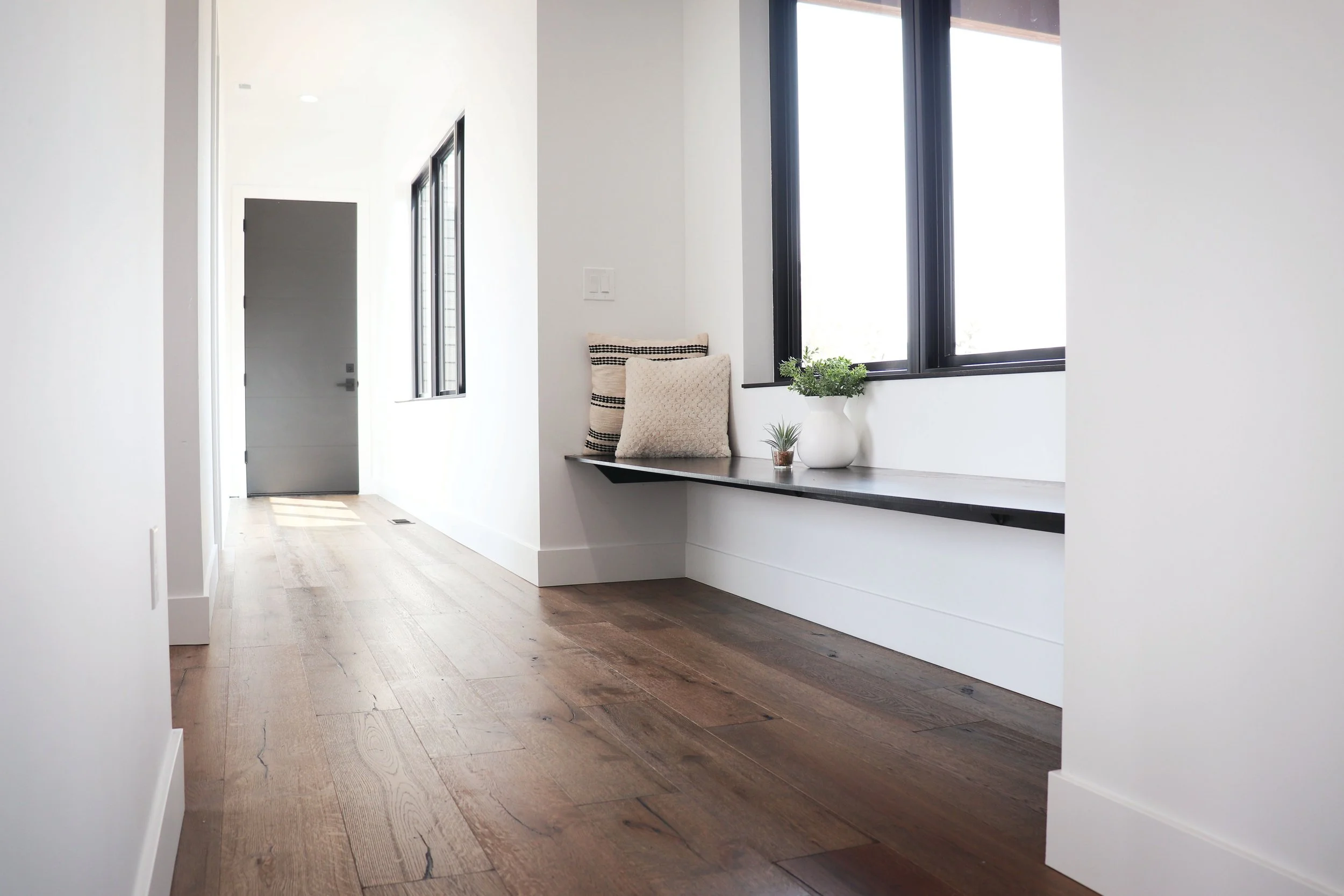 Bright hallway with white walls, wooden floor, and a window seat with cushions and potted plants.