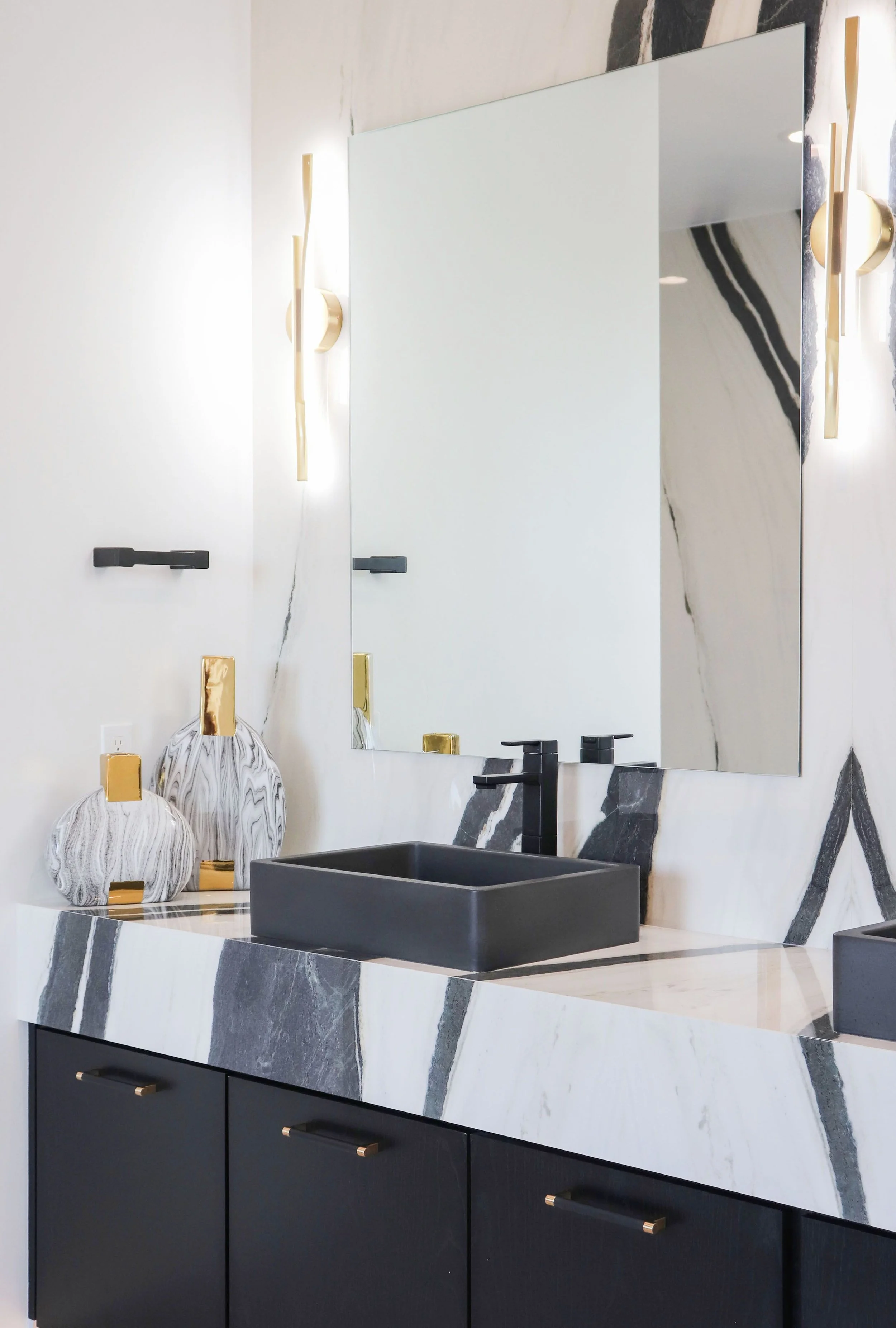 Modern bathroom vanity with a black rectangular sink, a large mirror, black cabinet with gold drawer handles, wall-mounted black faucet, decorative vases with gold accents, and contemporary wall sconces with gold and white design.