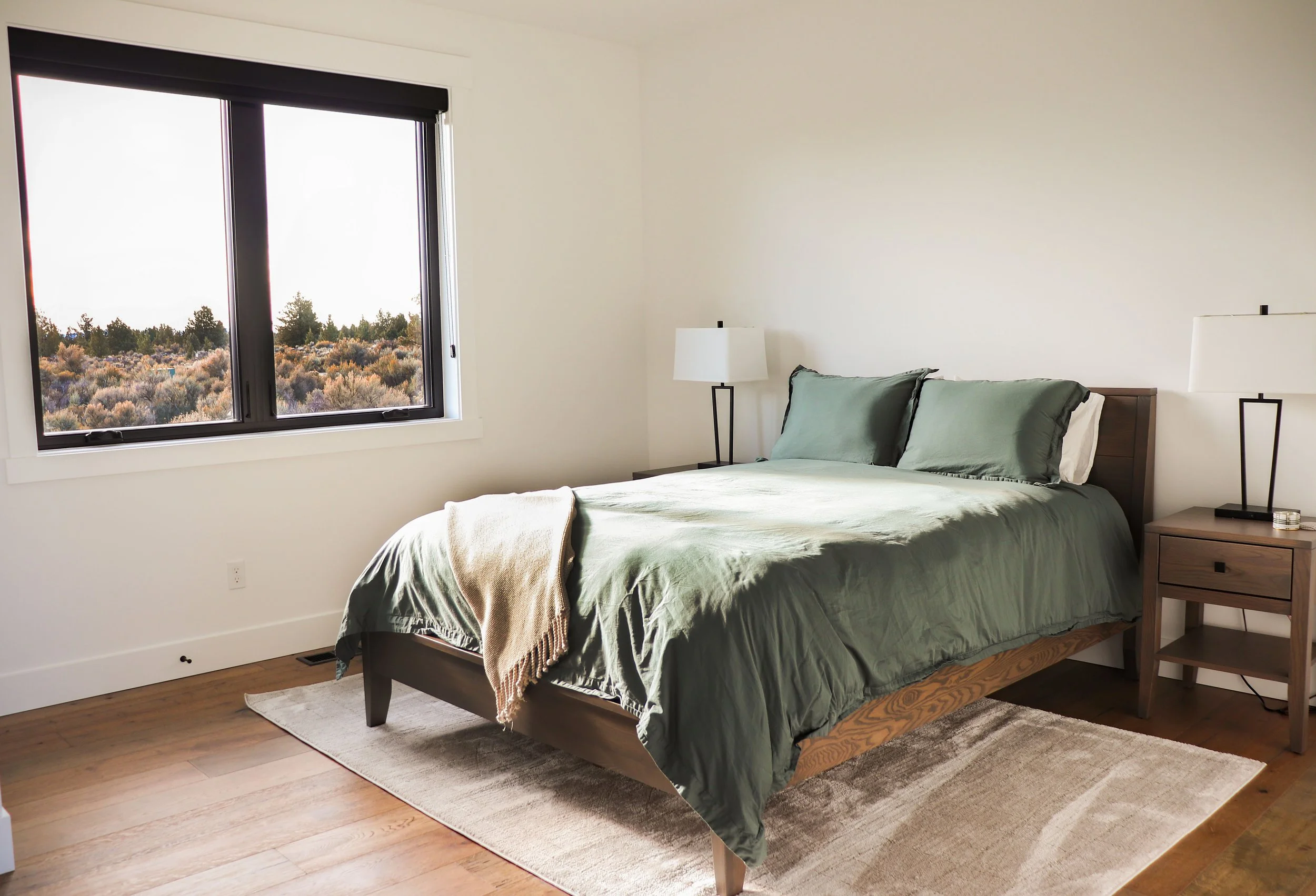 A bedroom with a large window showing a natural landscape outside, a bed with an olive-green duvet and pillows, wooden nightstands with white lamps, and a beige throw blanket on the bed.