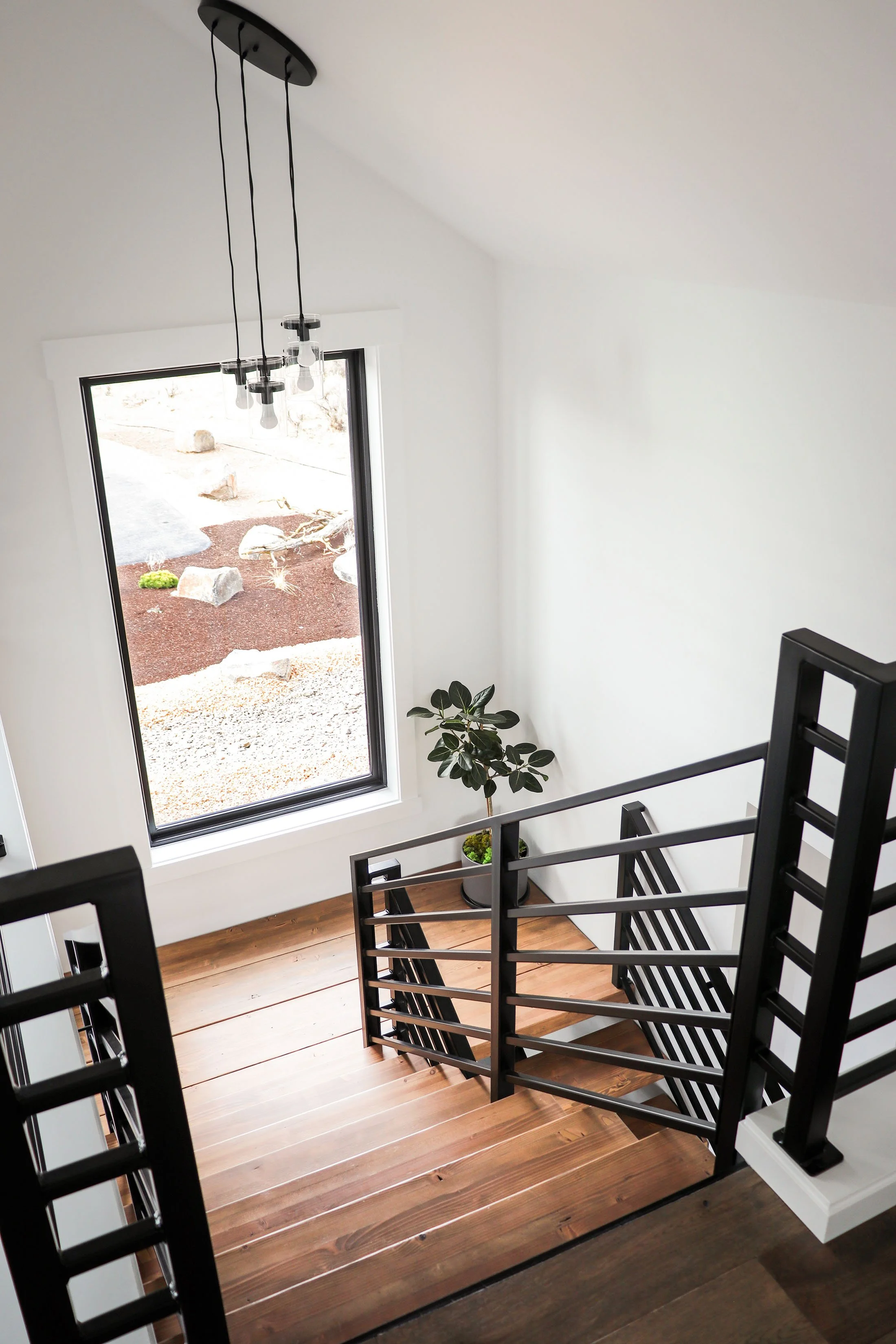 Indoor view of a staircase with black railings, a large window showing outdoor rocks and red mulch, and a black hanging light fixture with exposed bulbs.