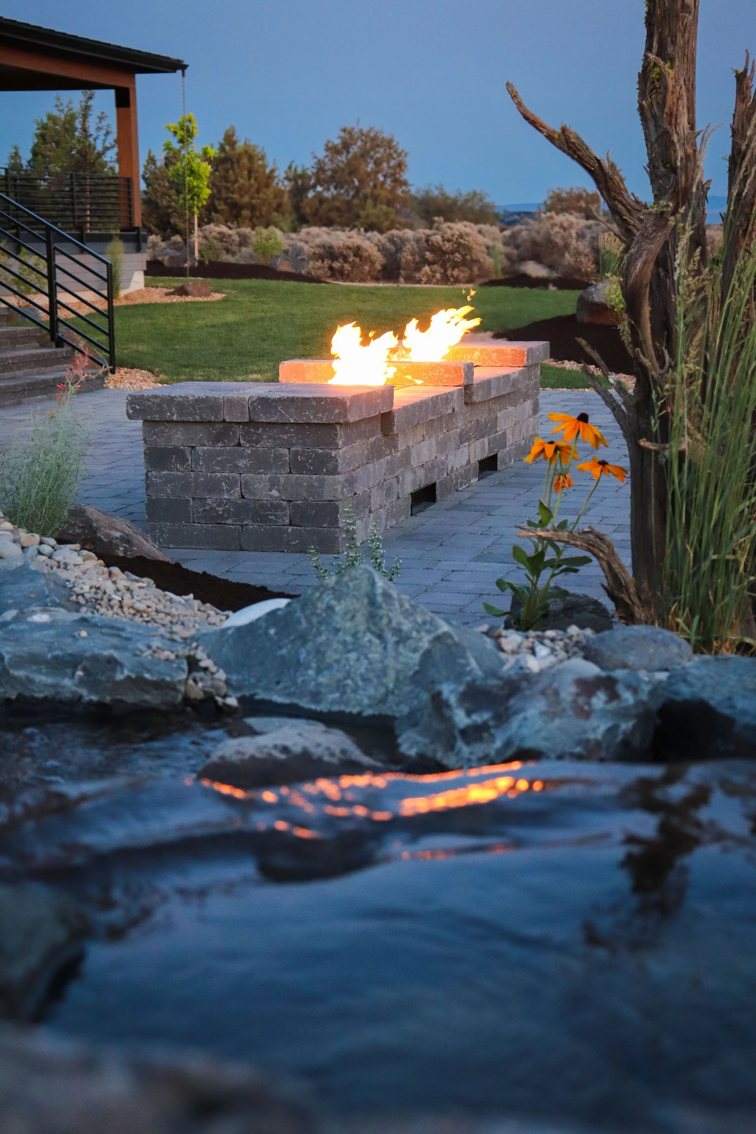 A backyard with a stone fire pit on a patio, surrounded by grass, trees, and plants, with a view of the evening sky.