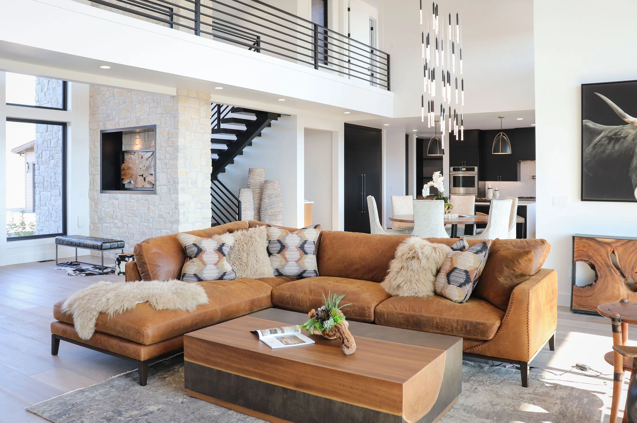 Modern open-concept living room with a brown sectional sofa adorned with patterned and furry pillows, a wooden coffee table with a plant arrangement, a large abstract painting of a bull, a stone accent wall with a built-in fireplace, and a staircase 