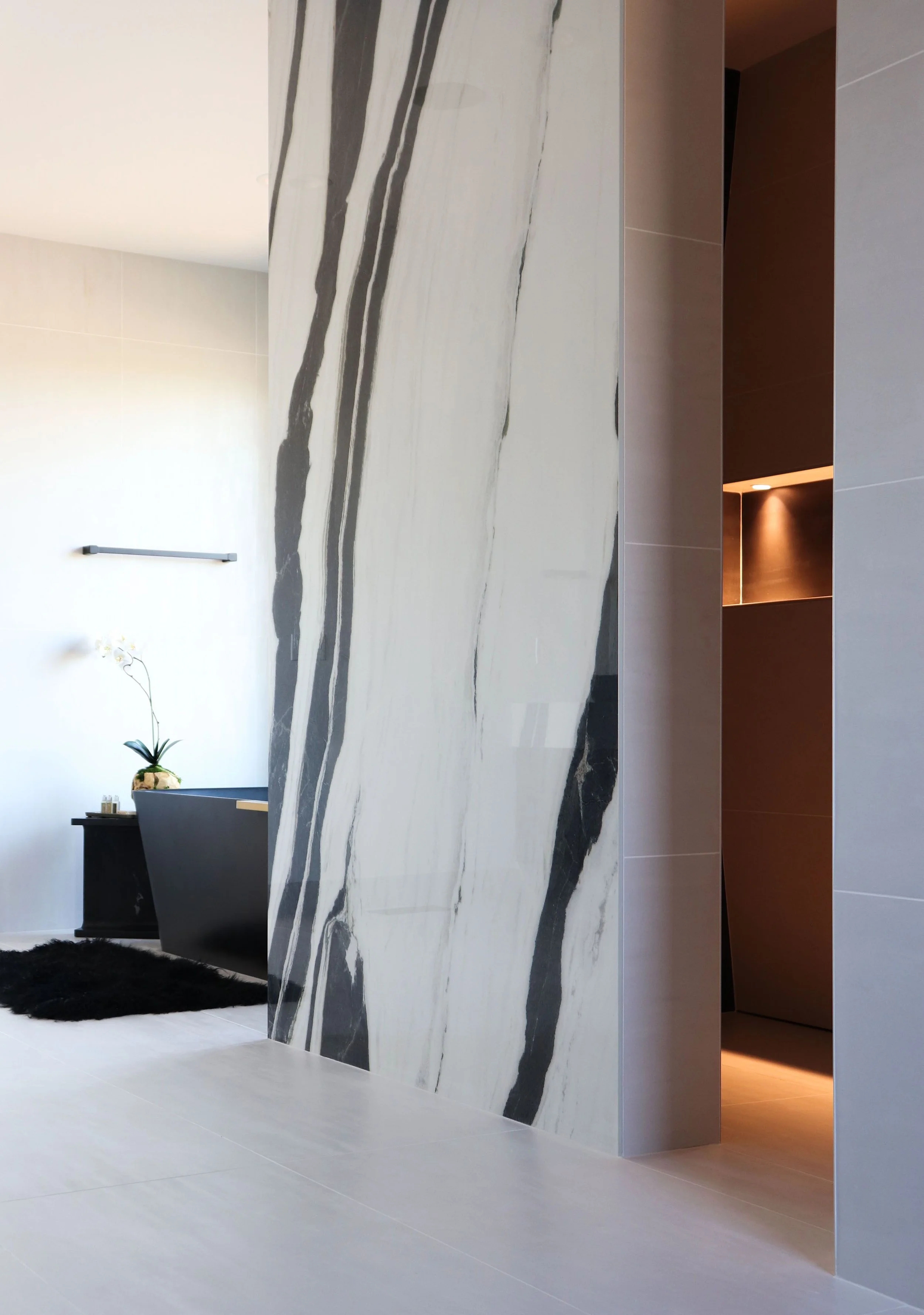 Living room with a black bathtub, a white orchid plant, black rug, and a large marble wall with black and white veining.