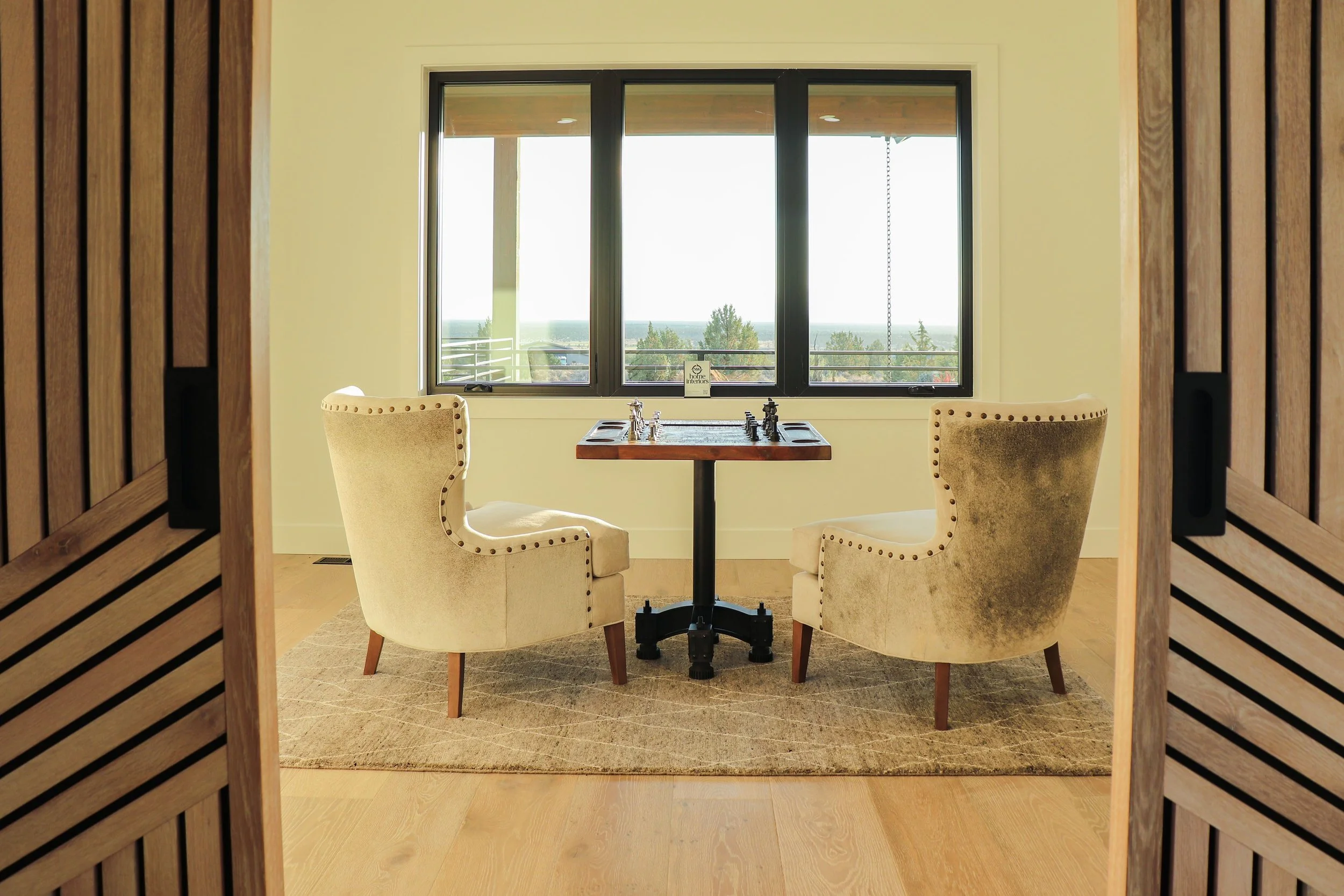 A room seen through a wooden sliding door, featuring two beige upholstered armchairs flanking a small wooden table with a chessboard, positioned in front of a large window with a view of trees and a balcony.