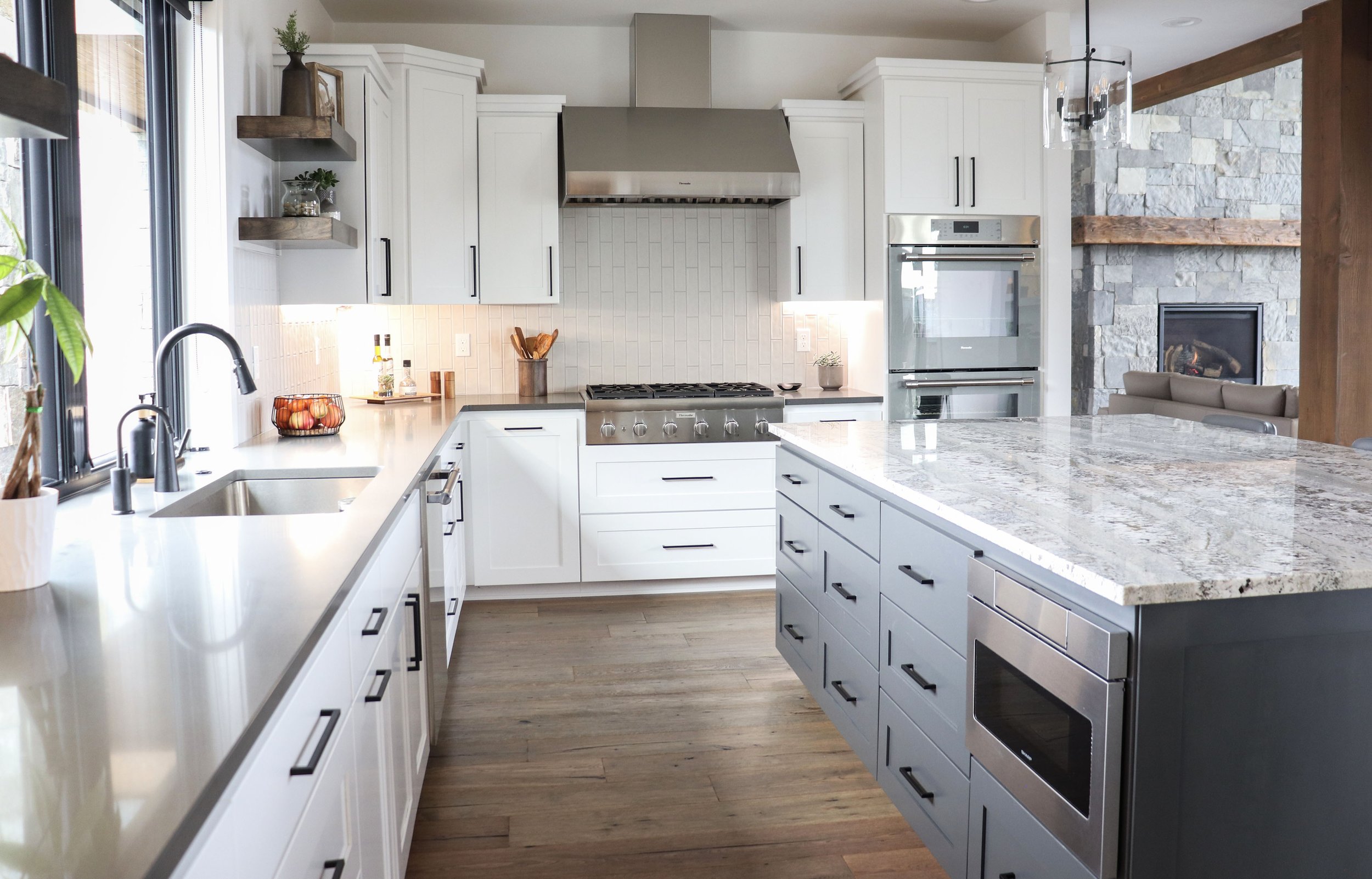 Modern kitchen with white cabinets, gray island, stainless steel appliances, stone fireplace, large window with plants, and hardwood floors.