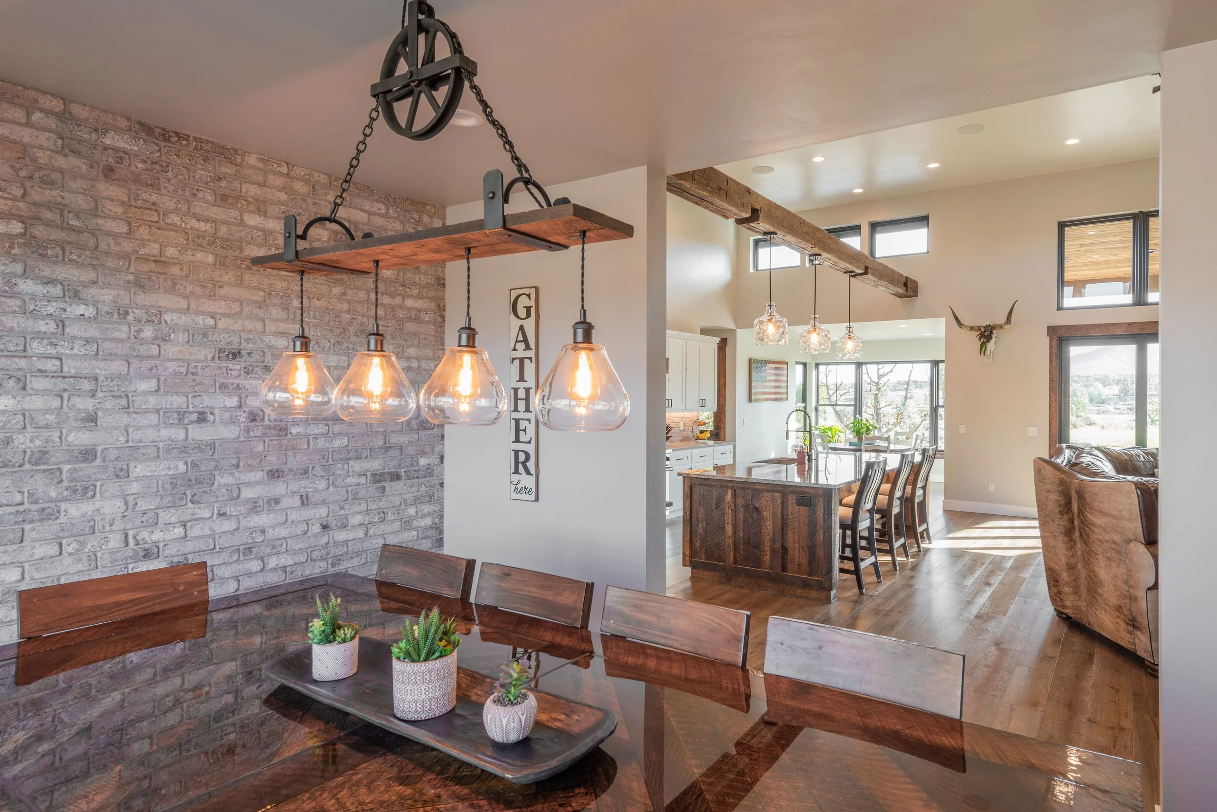 A dining area with a dark wood table, four matching chairs, and a modern chandelier with four glass pendant lights hanging from a wooden beam. The background shows an open kitchen with an island, barstools, and a living space with windows and outdoor