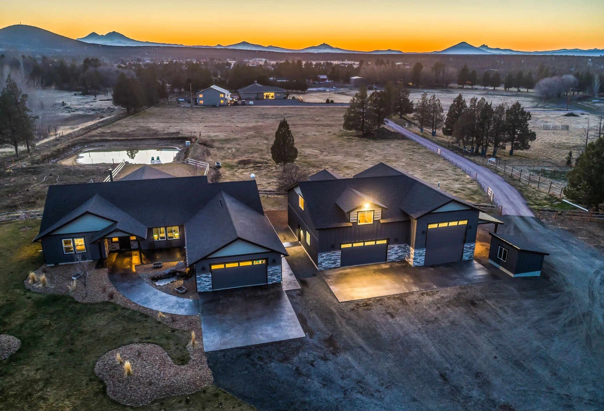 Aerial view of two modern houses with driveway and front yard, illuminated with outdoor lights during sunset, in a rural area with trees, a pond, and distant mountains.