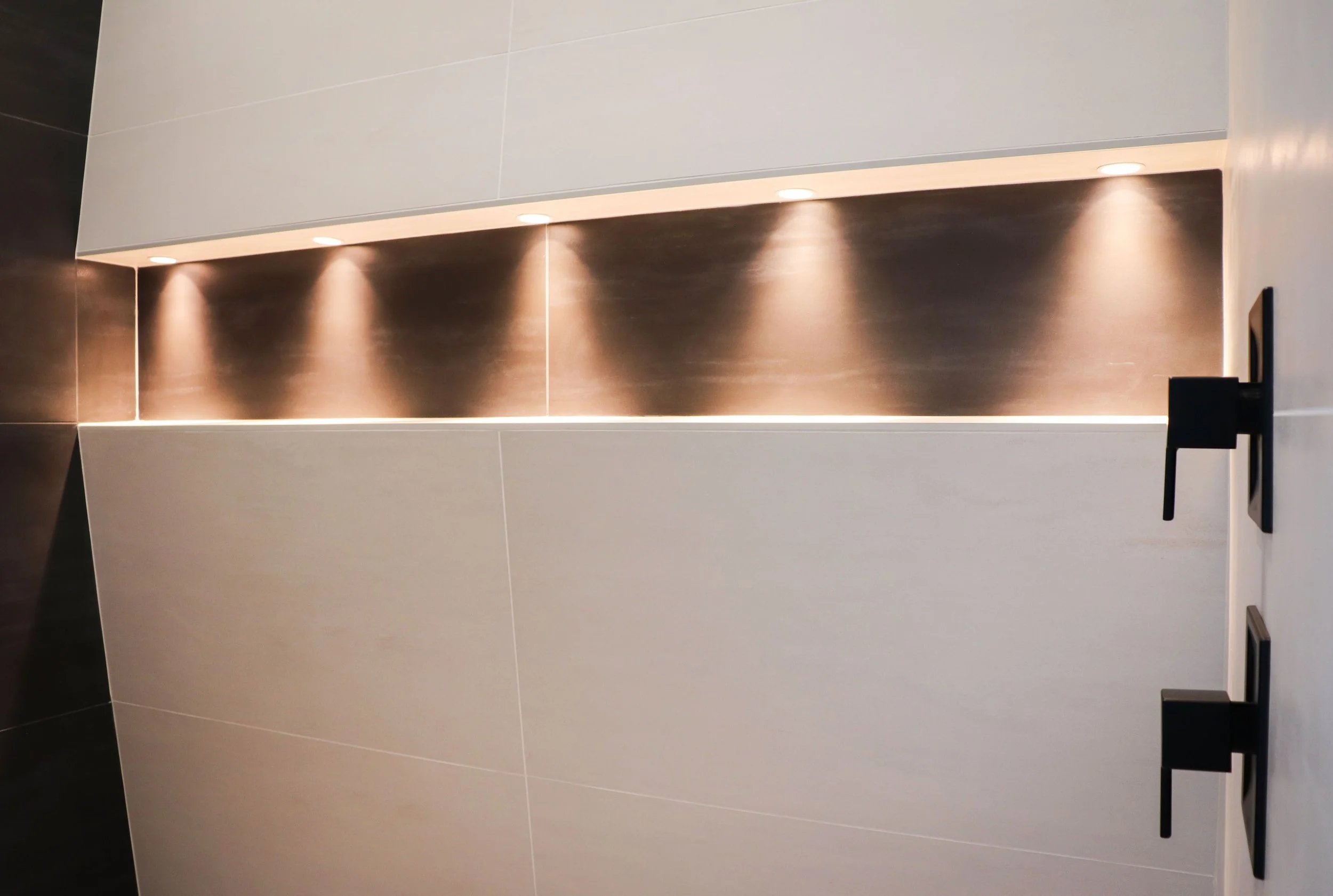 Close-up of a modern bathroom wall with dark and white tiles, under-cabinet lighting, and black handles.