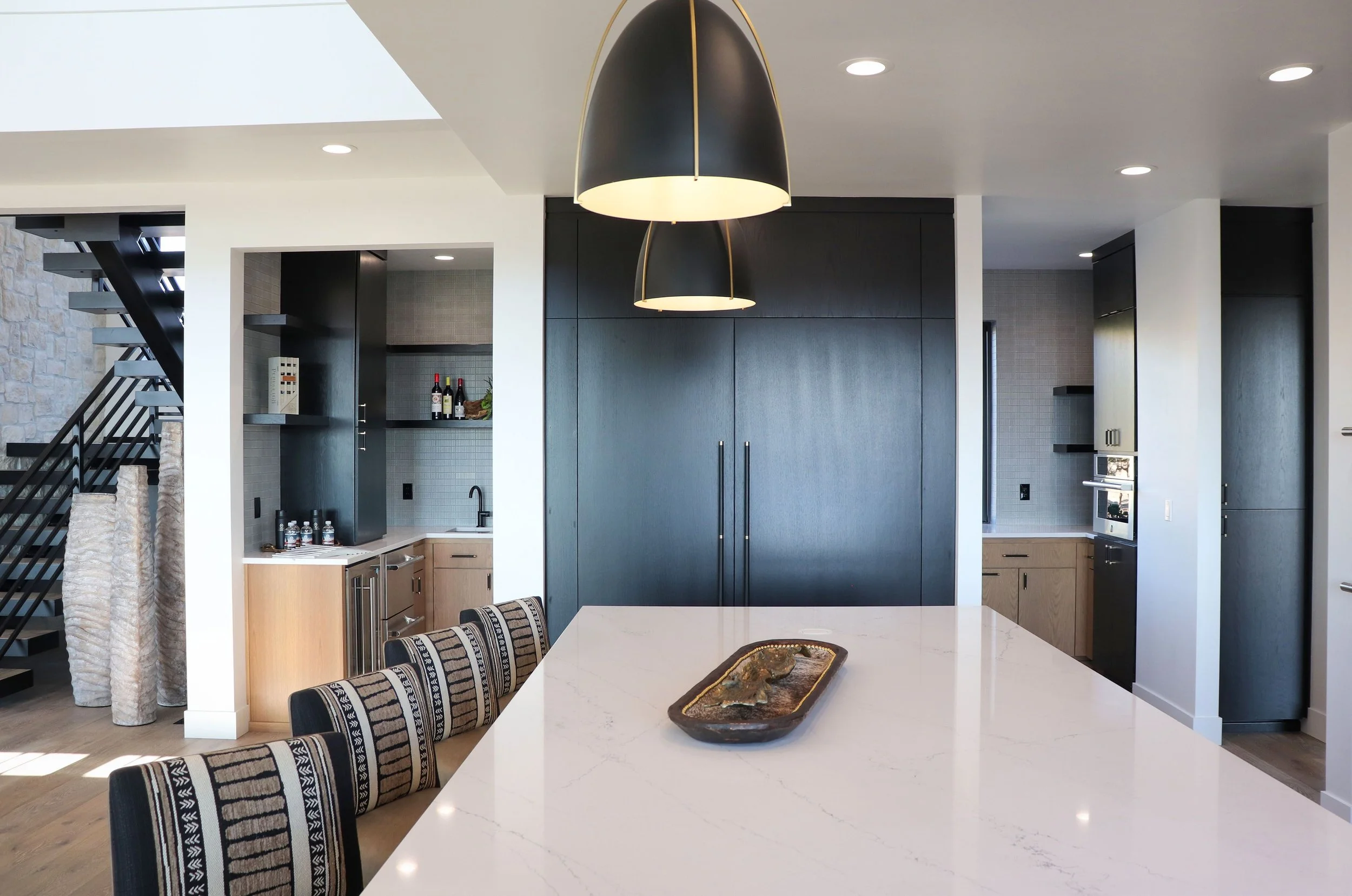 Modern kitchen with black cabinetry, white marble island, black pendant lights, and patterned dining chairs.