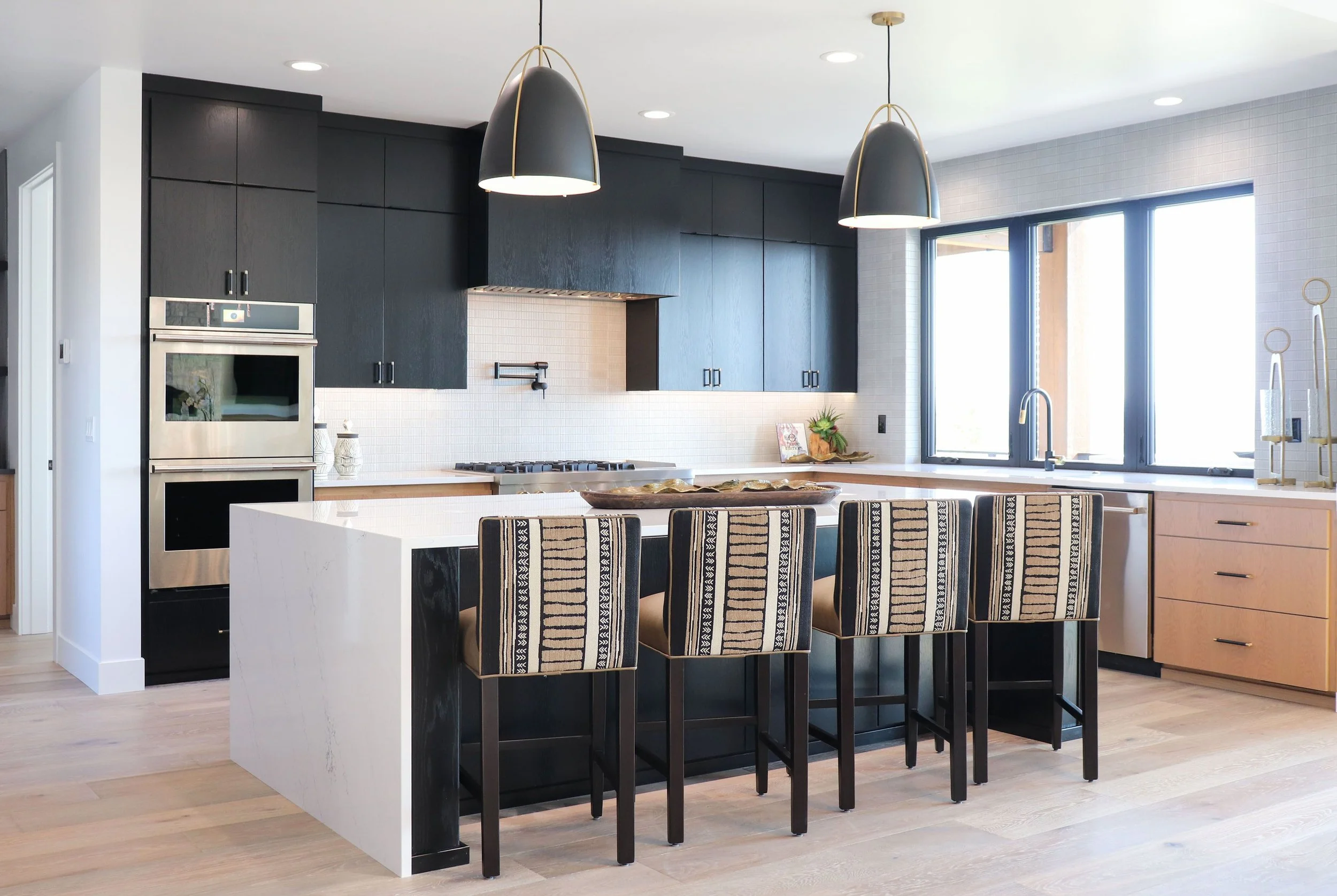 Modern kitchen with black upper cabinets, light wood lower cabinets, a large white island with a black side, bar stools with patterned fabric, stainless steel oven, and large windows letting in natural light.