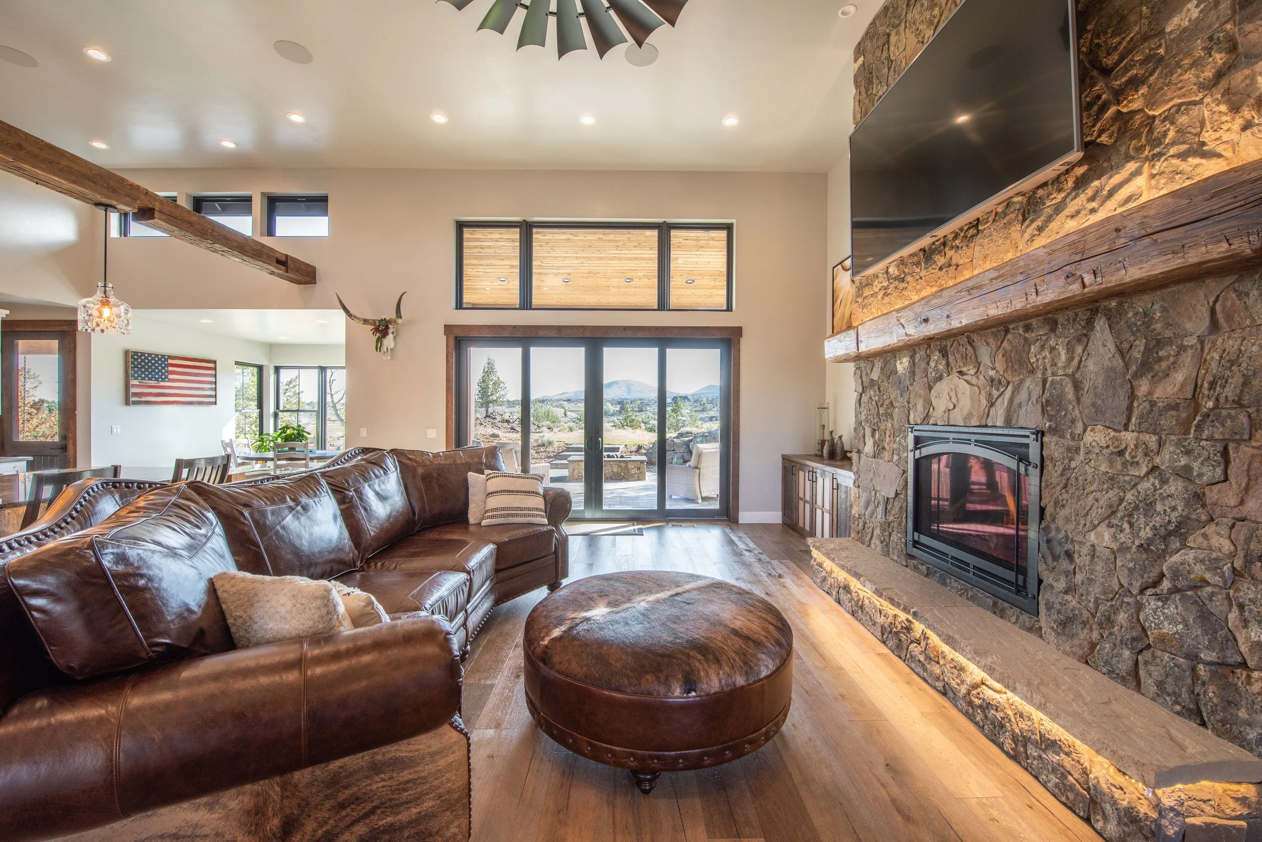 Living room with large stone fireplace, wooden beam, leather sectional sofa, and a round leather ottoman, with sliding doors leading to outdoor patio and views of mountains.