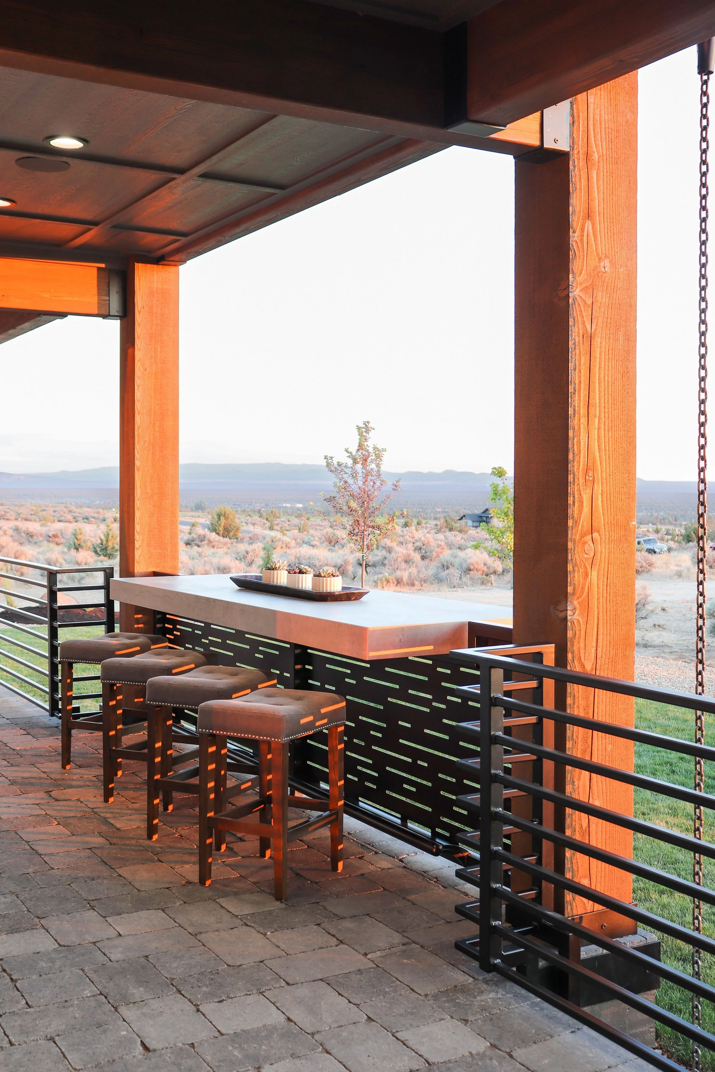 Outdoor patio with four bar stools at a bar-height counter, overlooking a scenic landscape with distant mountains and open fields.
