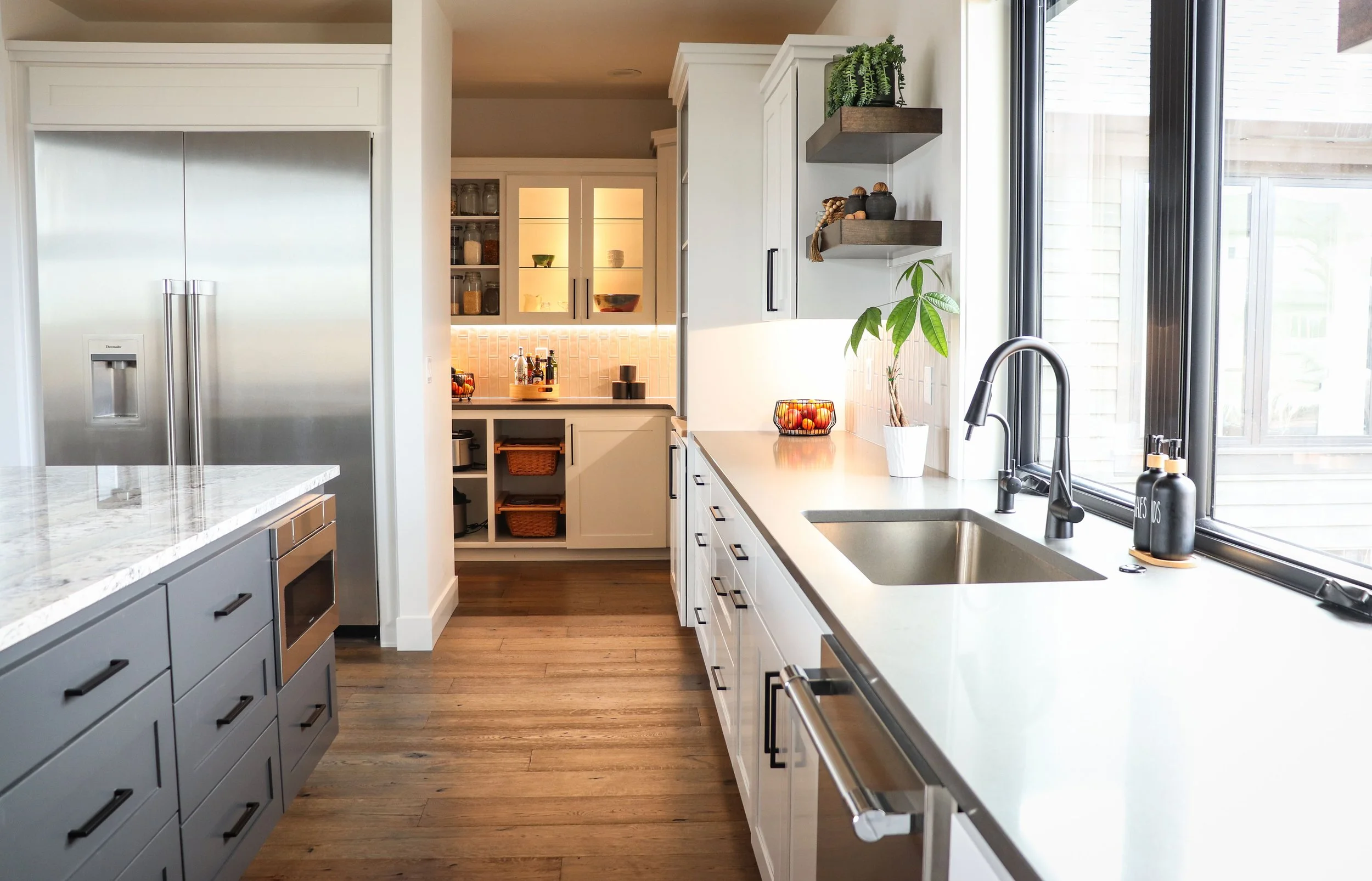 Modern kitchen with white and gray cabinetry, stainless steel refrigerator, black faucet, white countertops, and wooden floor, with a large window above the sink.