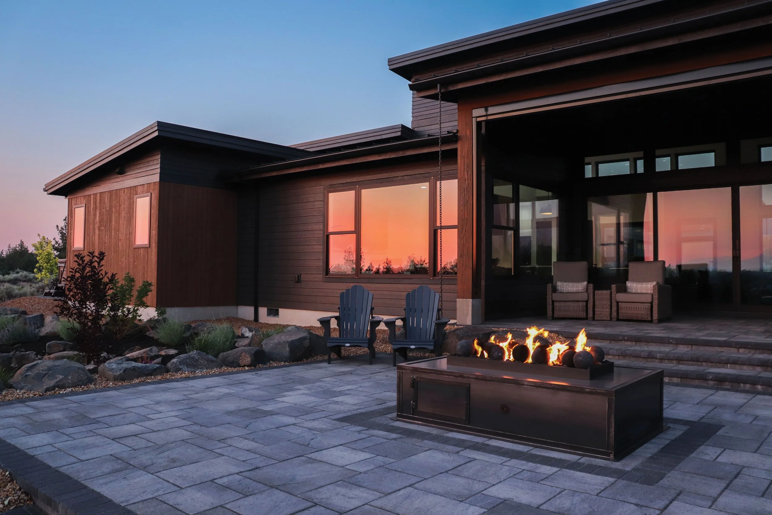 A modern house with dark wood exterior windows reflecting a sunset sky, a paved patio with two blue Adirondack chairs, two armchairs on the covered porch, and a rectangular firepit with flames, surrounded by a landscaped yard with rocks and plants.
