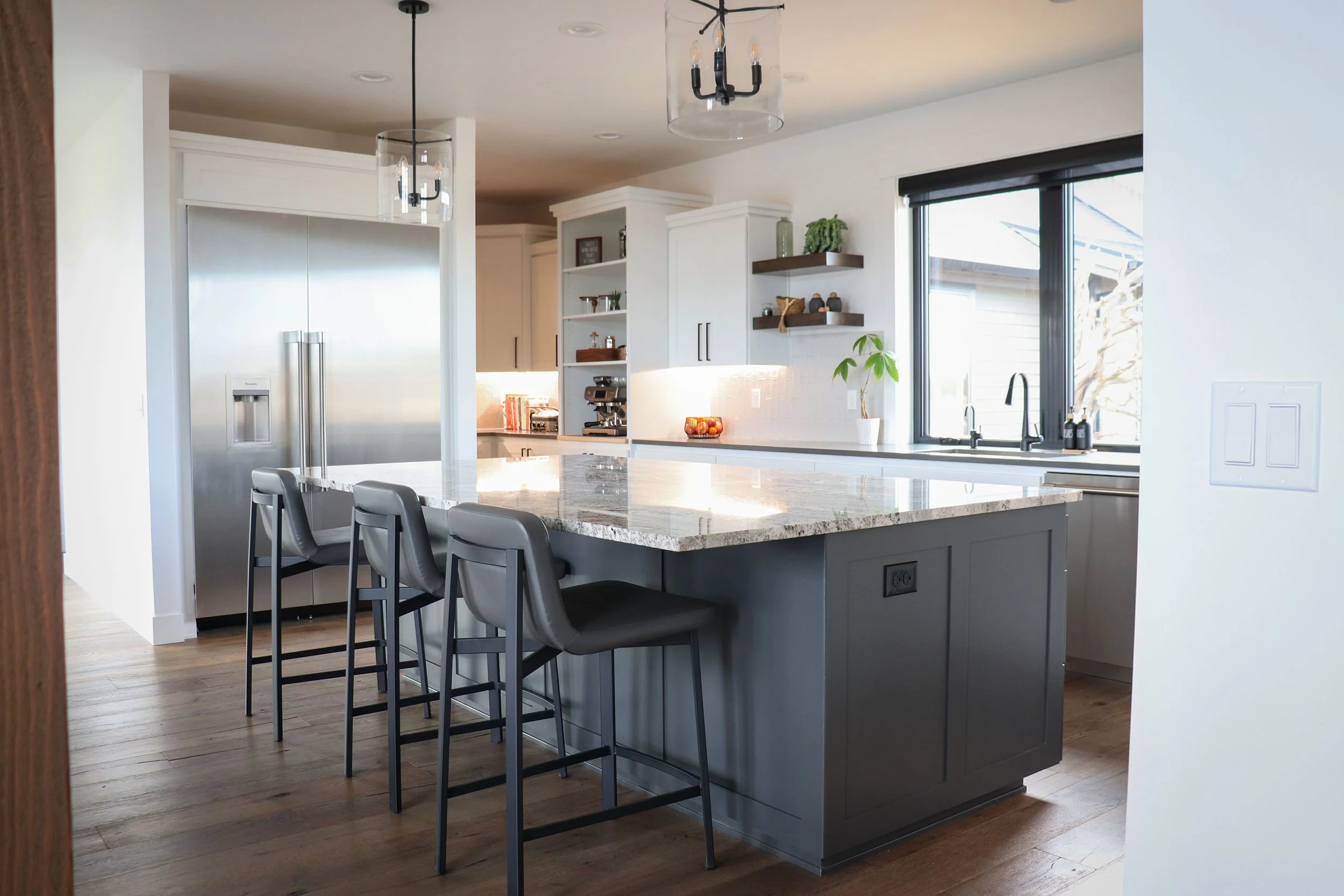 Modern kitchen with a large granite island, three gray barstools, stainless steel refrigerator, white cabinets, open shelving, and a window with black blinds.