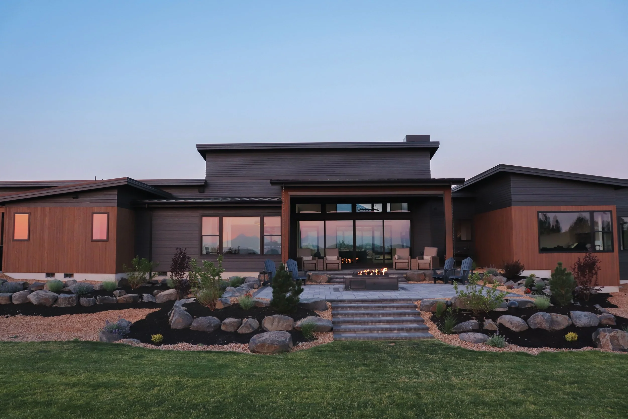 Modern house with a landscaped garden, patio seating area, and firepit, overlooking a mountain view at dusk.